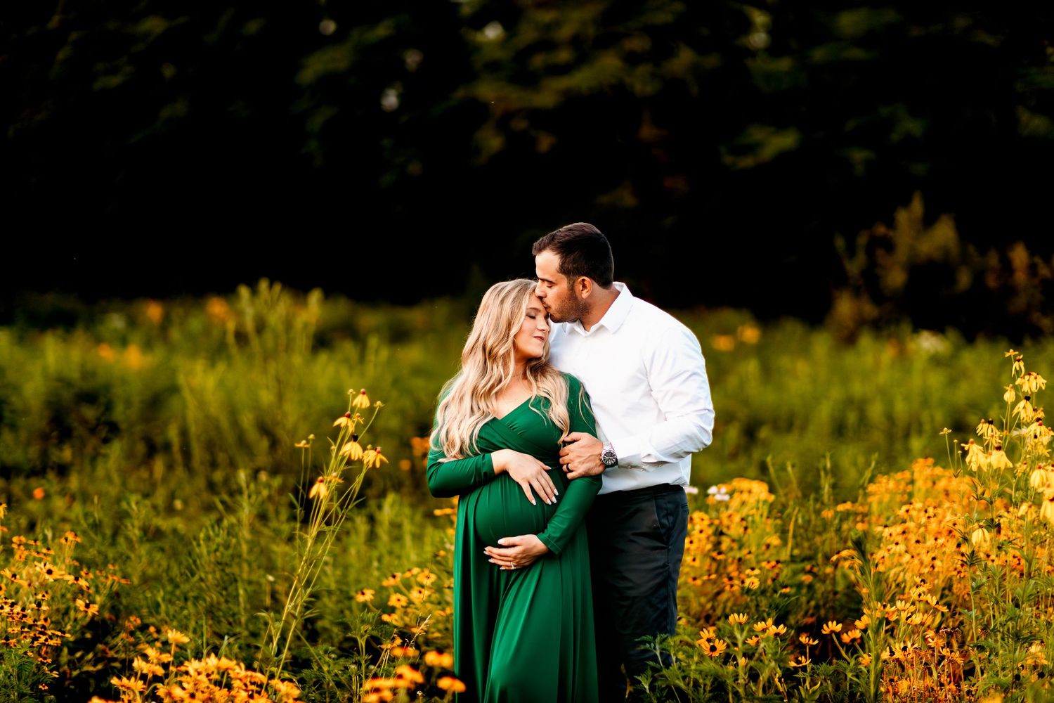 A couple stands in a lush field, surrounded by yellow flowers. The woman, in a green dress, is pregnant and embraced by her partner
