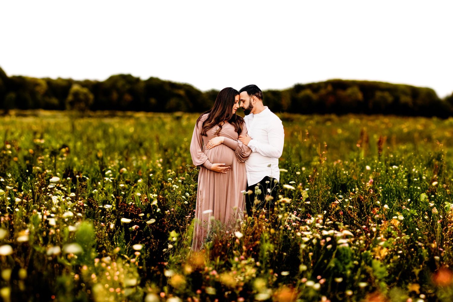 A couple stands closely in a lush field of wildflowers, the woman pregnant, wearing a flowing dress. The setting is serene, conveying love