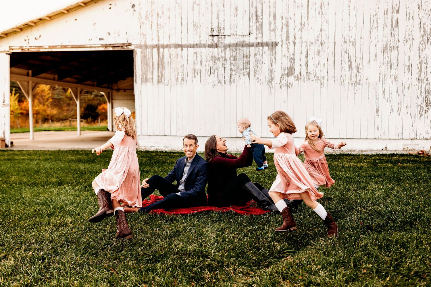 Family sitting on a picnic blanket in front of a rustic barn, their three young daughters in pink dresses joyfully dancing around them