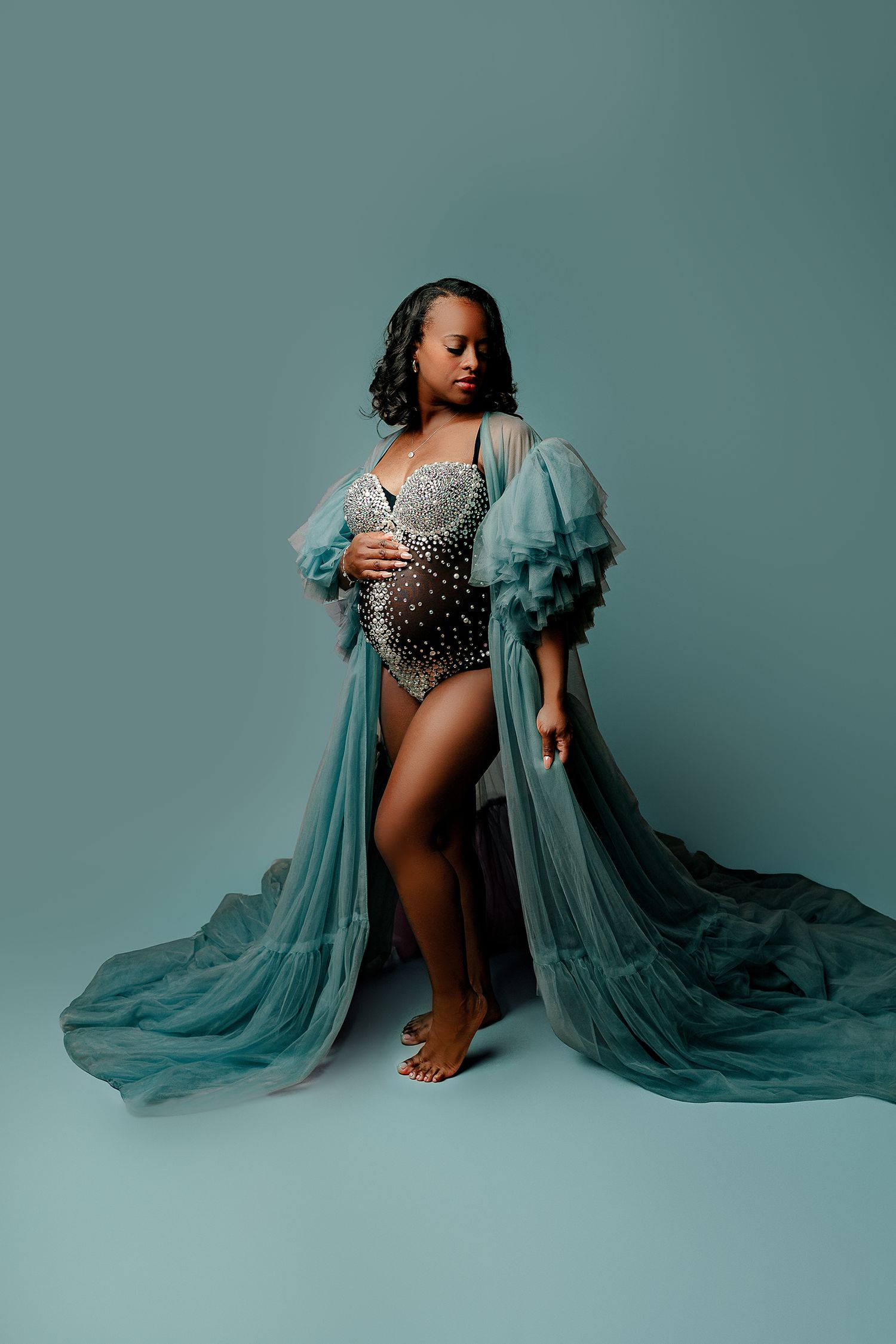 Pregnant woman in elegant, flowing teal robe over embellished bodysuit stands barefoot against a matching backdrop, exuding grace