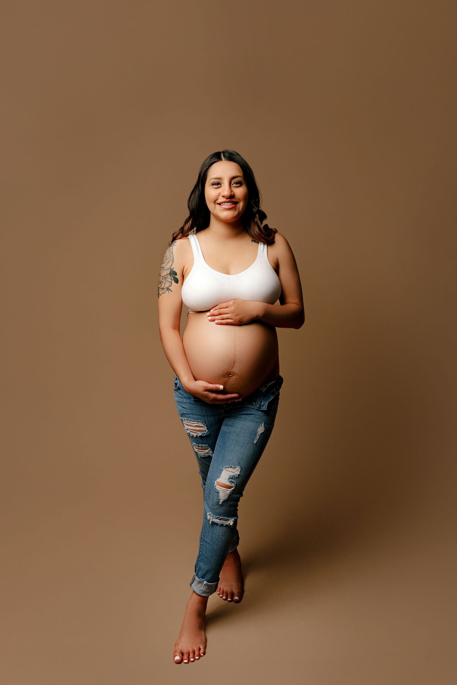 Pregnant woman smiling, standing barefoot against a plain brown background. She wears a white top and ripped jeans