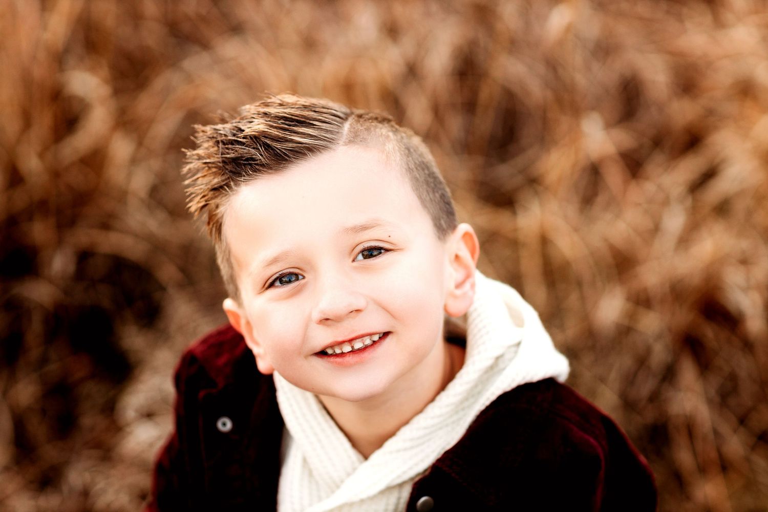 A smiling young boy with short hair wearing a dark jacket and white sweater sits in front of a blurred, earthy background