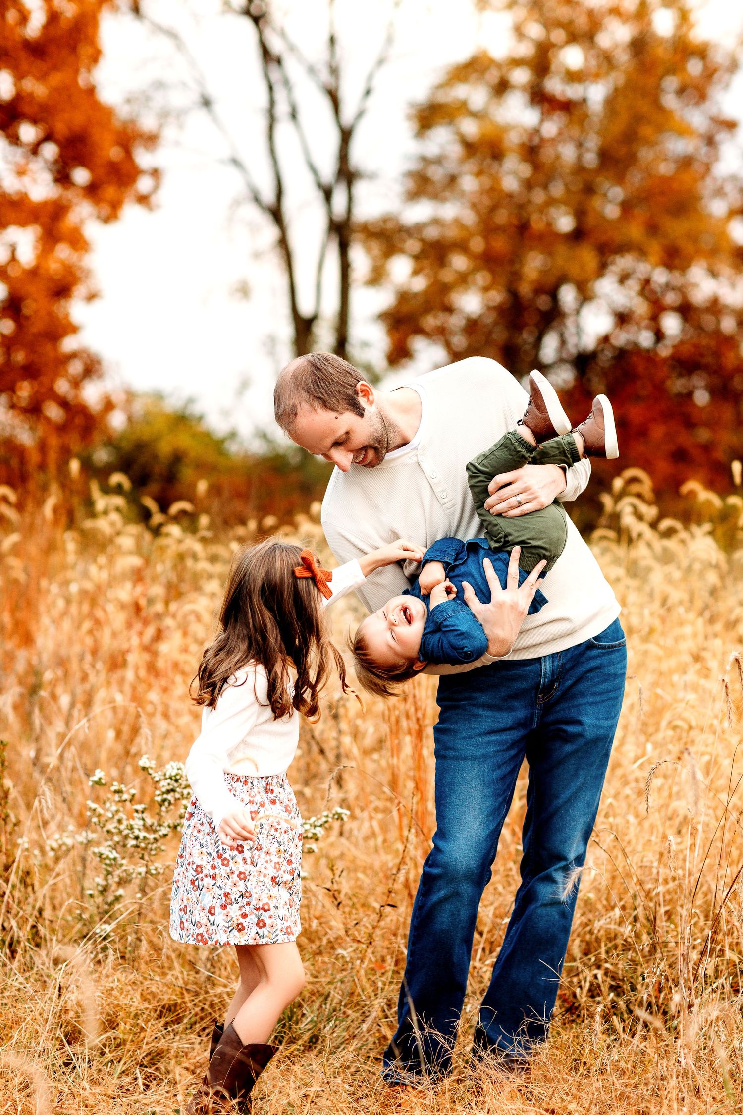 A father playfully holds a young boy upside down in an autumn field. A girl stands nearby, smiling.