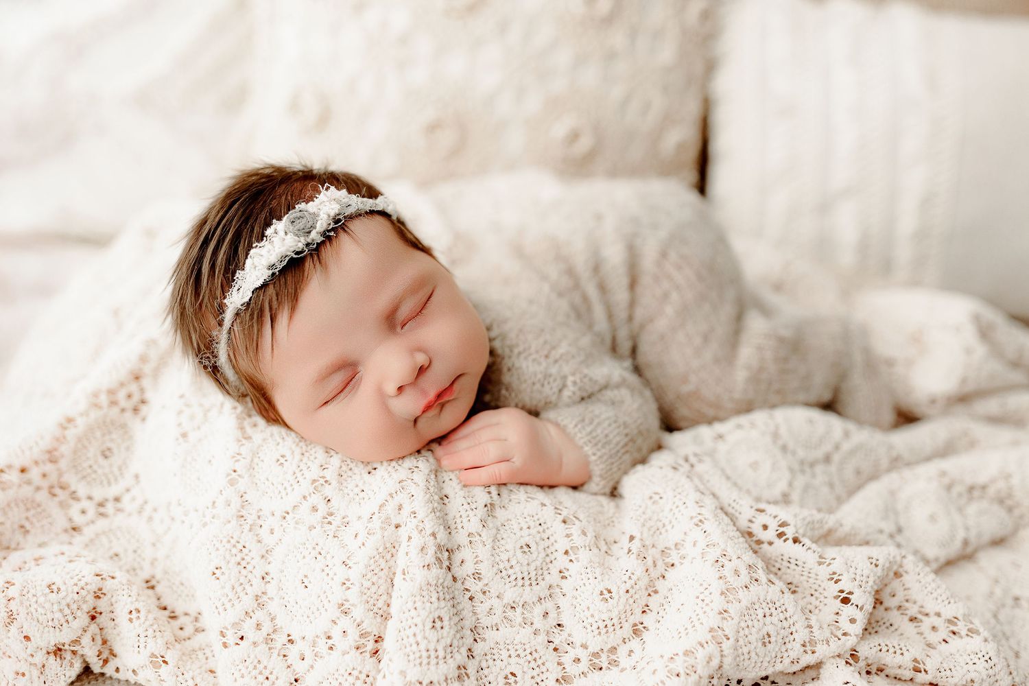 A peaceful baby sleeps on a cream lace blanket, wearing a soft white outfit and a delicate headband with flowers. The scene is cozy