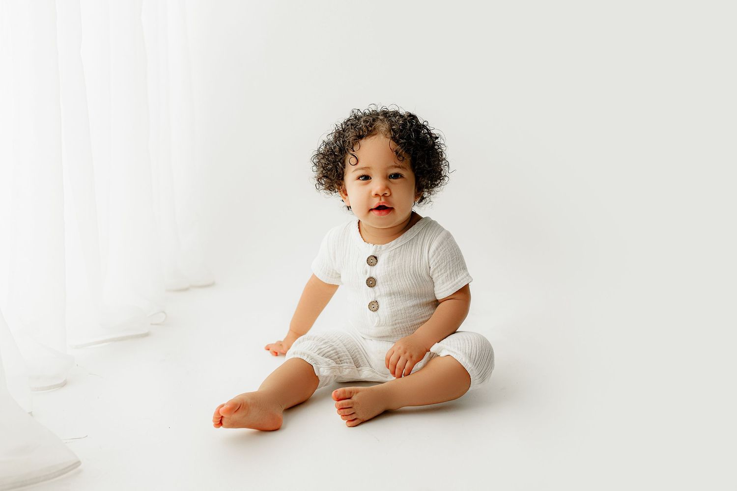 A toddler with curly hair sits on a white floor, wearing a white outfit with big buttons. The backdrop is minimalistic