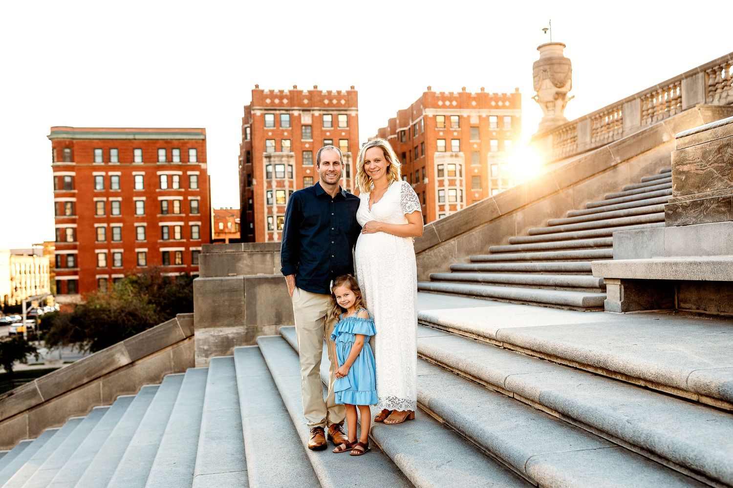 A family poses on steps at sunset at Indiana War Memorial, with brick buildings in the background. The man, woman, and child smile warmly