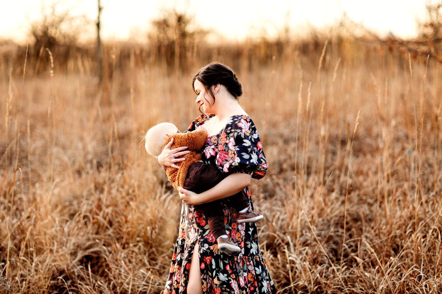 A woman in a floral dress lovingly holds a child in a peaceful field of tall, dry grass during a warm, golden sunset