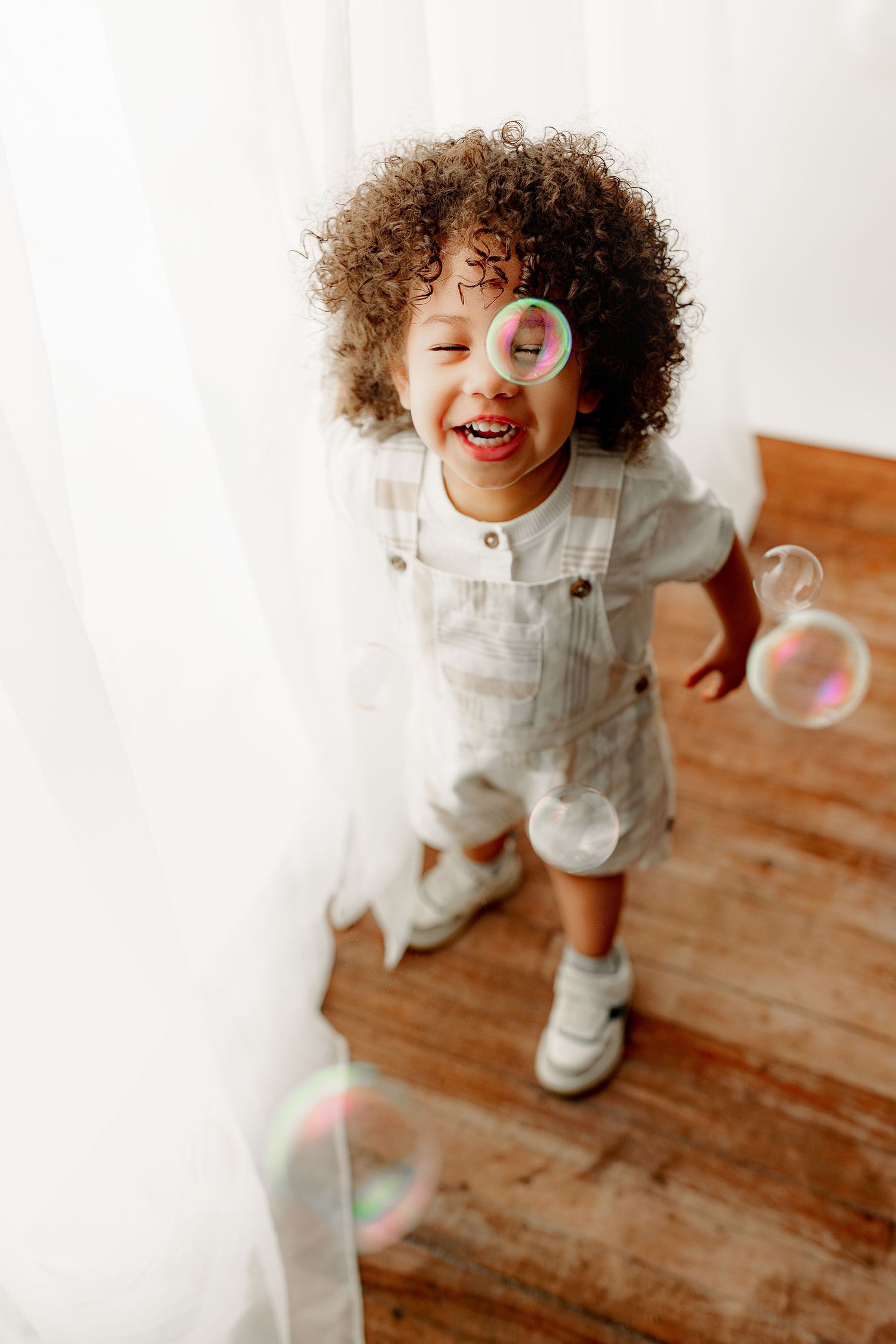 A joyful child with curly hair and overalls gleefully plays with bubbles in Central Indiana photography studio