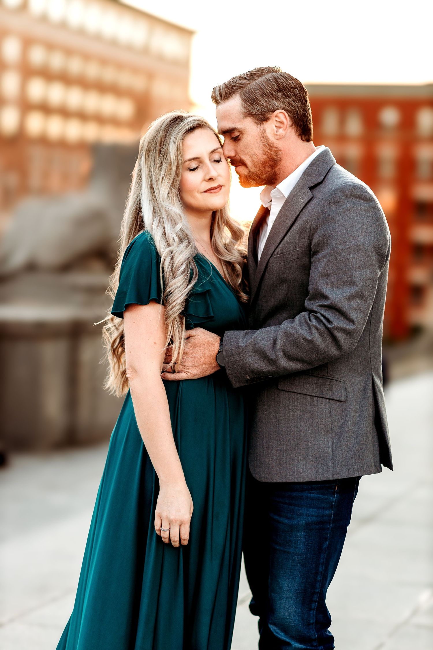 A couple stands embracing with sunlit city scene The woman in a teal dress has long hair, and the man in a gray jacket gently kisses her