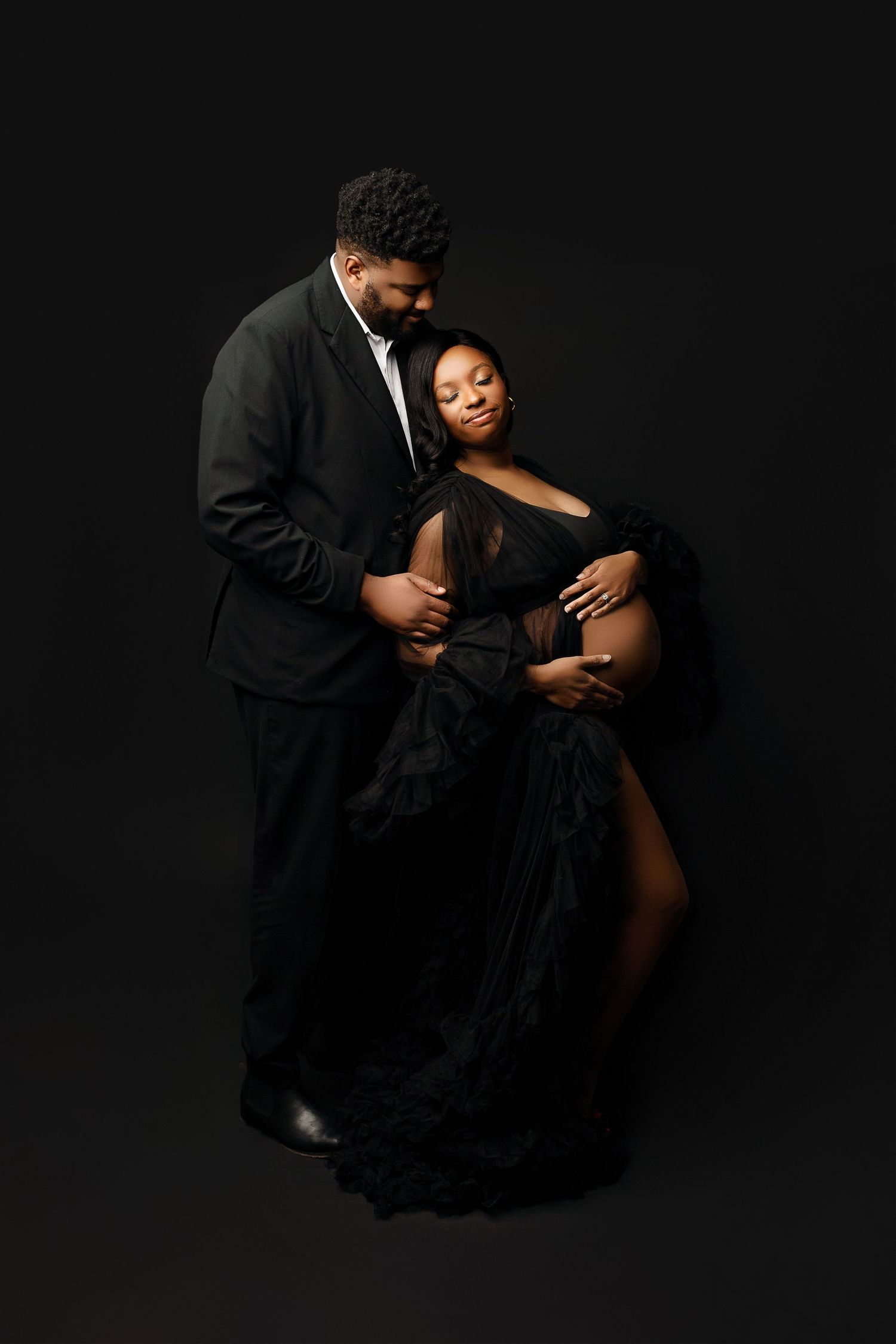 A couple poses intimately against a dark backdrop. The man wears a black suit, while the woman cradles her pregnant belly, dressed in black