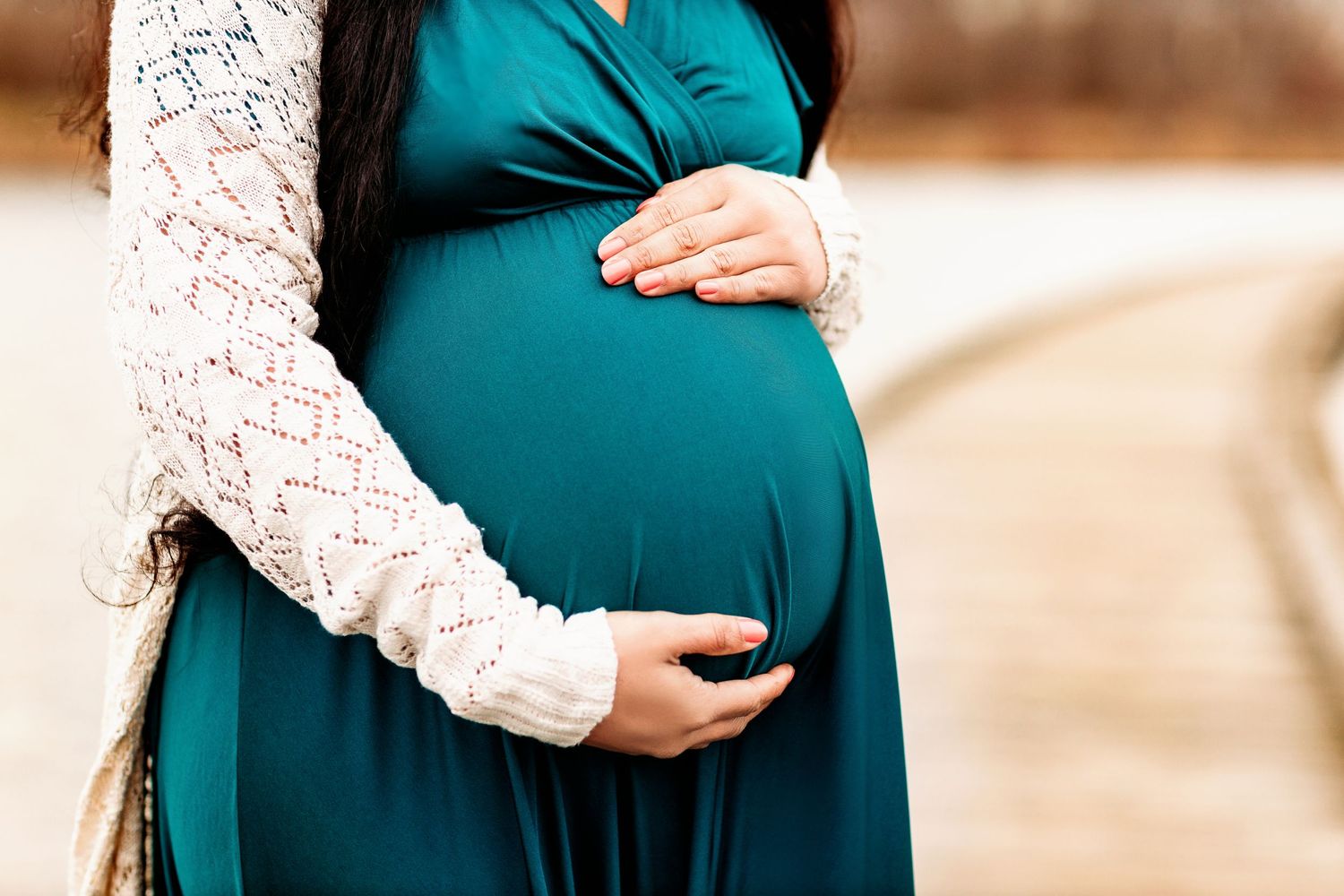 A pregnant woman in a teal dress cradles her belly with both hands. She wears a white knitted cardigan, standing on a blurred outdoor path