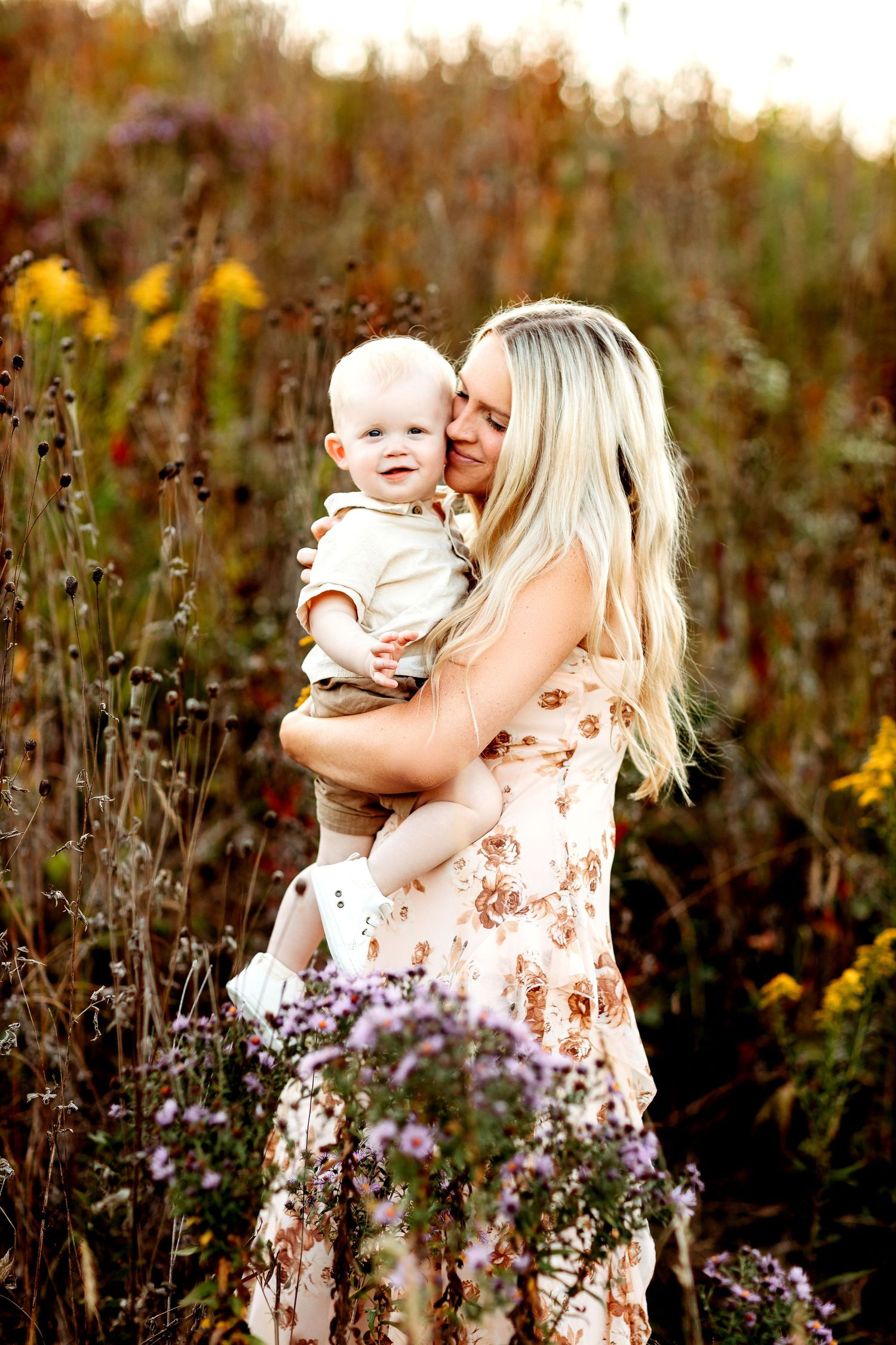 A woman with long blonde hair in a floral dress holds a smiling baby in a field of wildflowers