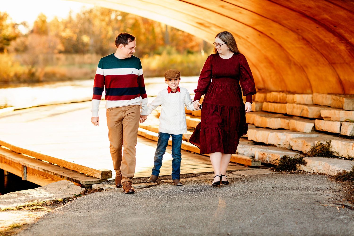 A family of three walks hand in hand under a wooden bridge in autumn. They wear fall colors, smiling warmly, with trees in the background