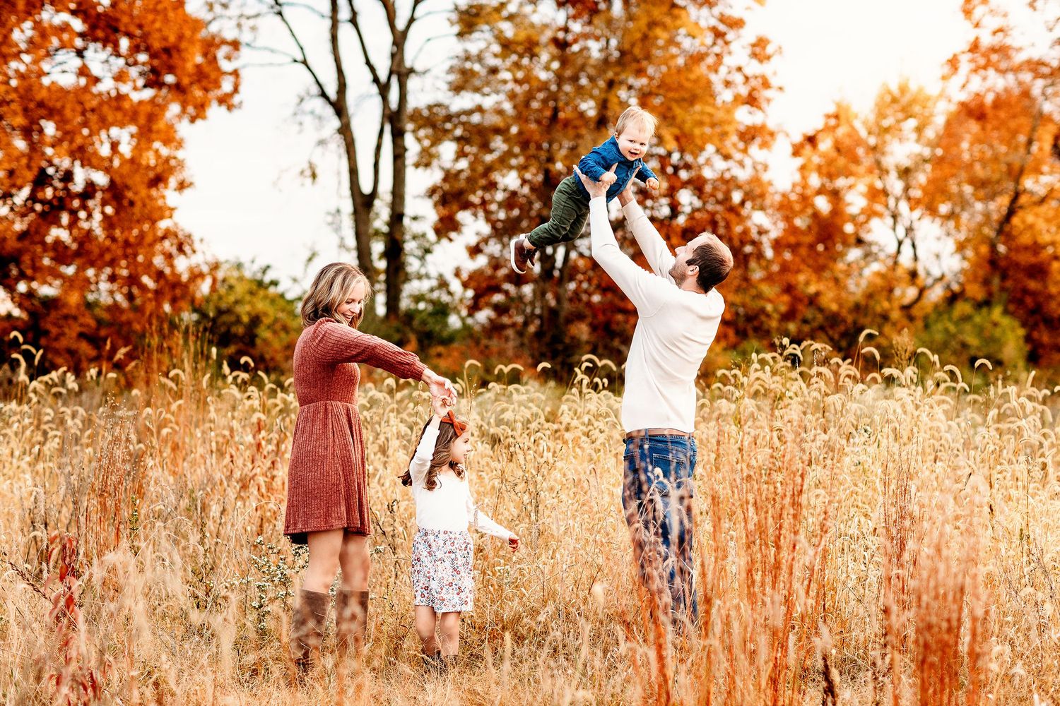 A joyous family in an autumn field. A man lifts a child high, while a woman and girl hold hands.
