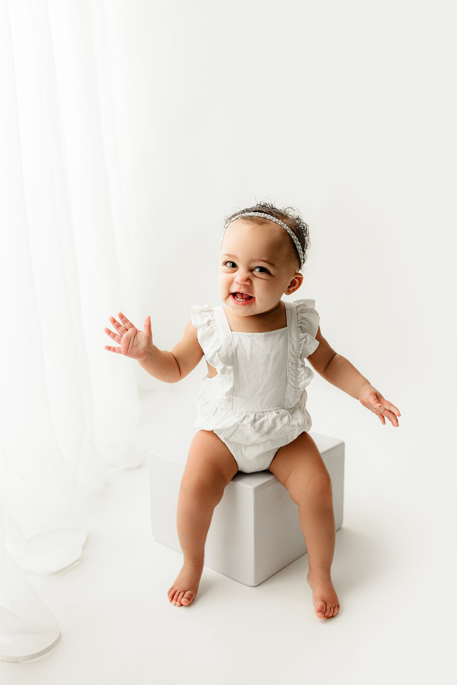 A smiling toddler in a white ruffled outfit sits on a white cube against a plain background. The child waves, creating a joyful tone