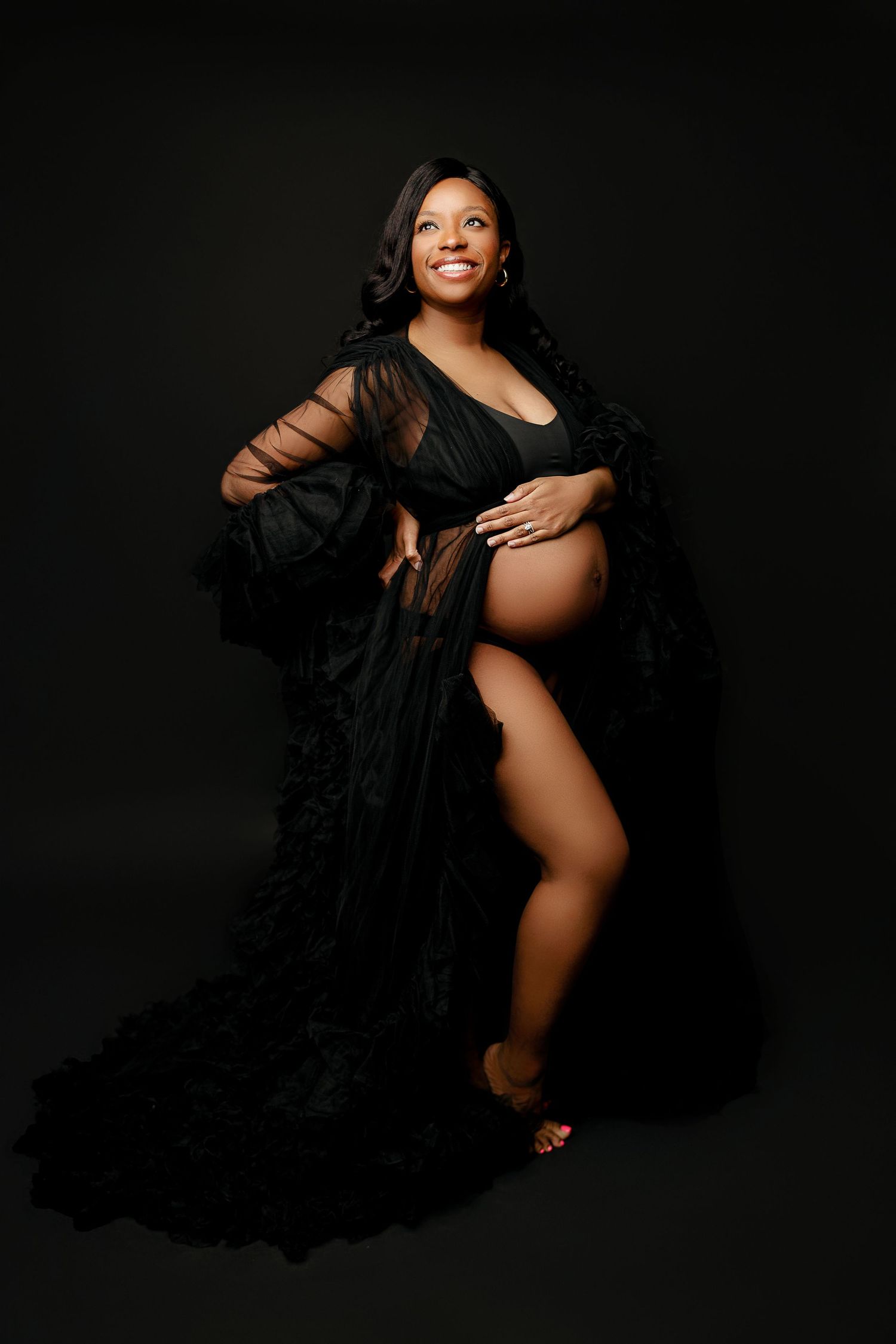 A pregnant woman in a black flowing gown stands against a dark background, smiling confidently. Her hand rests on her belly,