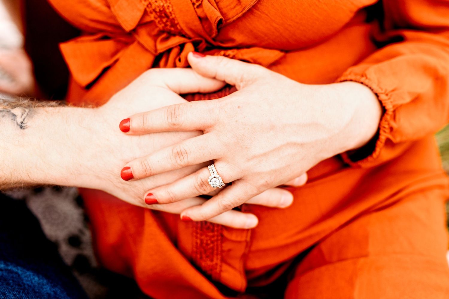 Close-up of a pregnant woman's hands resting on her belly, wearing an orange dress. Another person's hands gently hold hers
