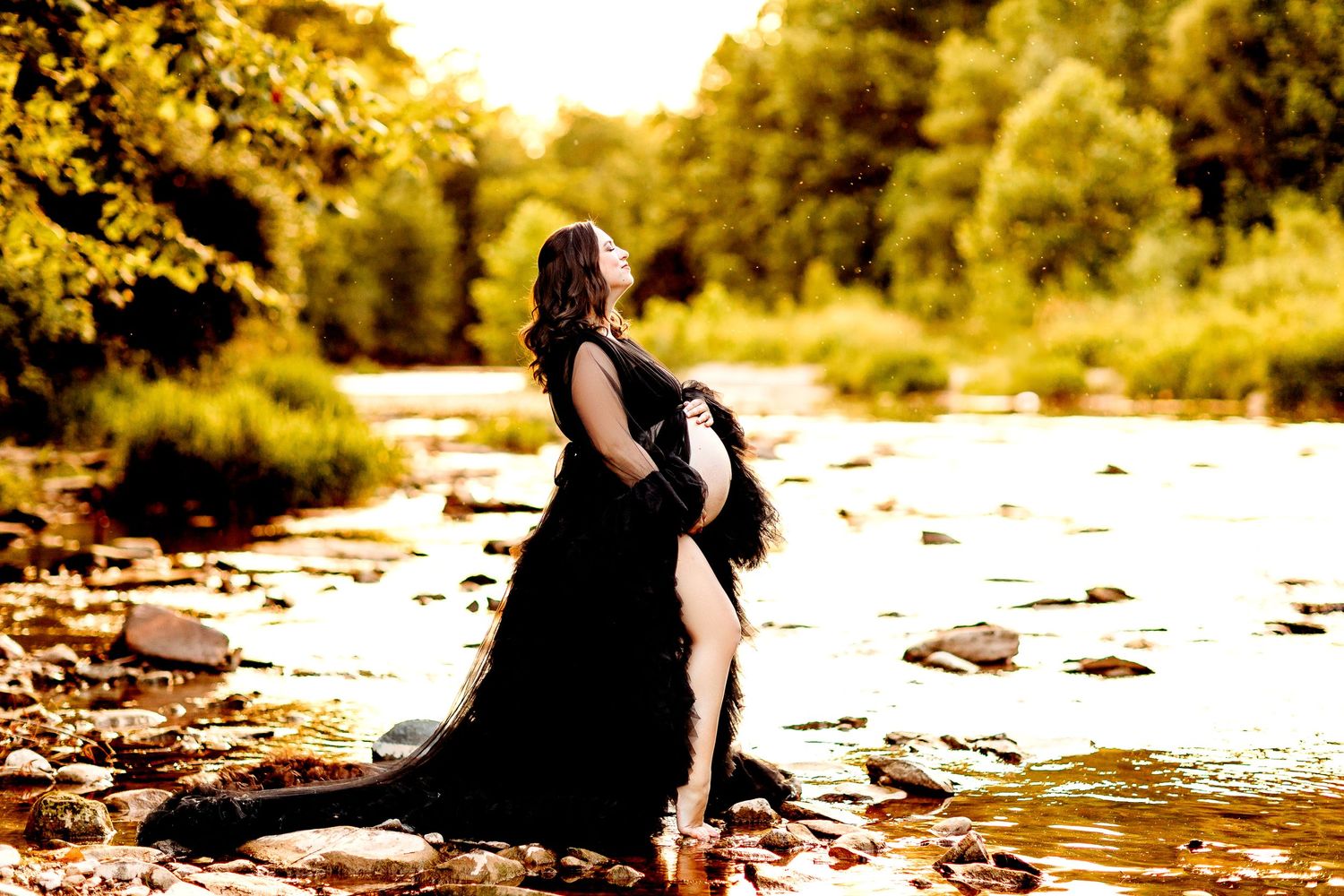 A pregnant woman in a flowing black dress stands on a rocky riverbank, looking up serenely. Sunlight filters through the trees