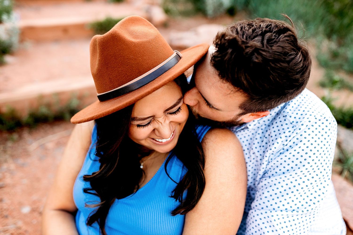 A smiling couple shares a tender moment outdoors. The man, in a blue shirt, kisses the woman's cheek. She wears a brown hat and blue top
