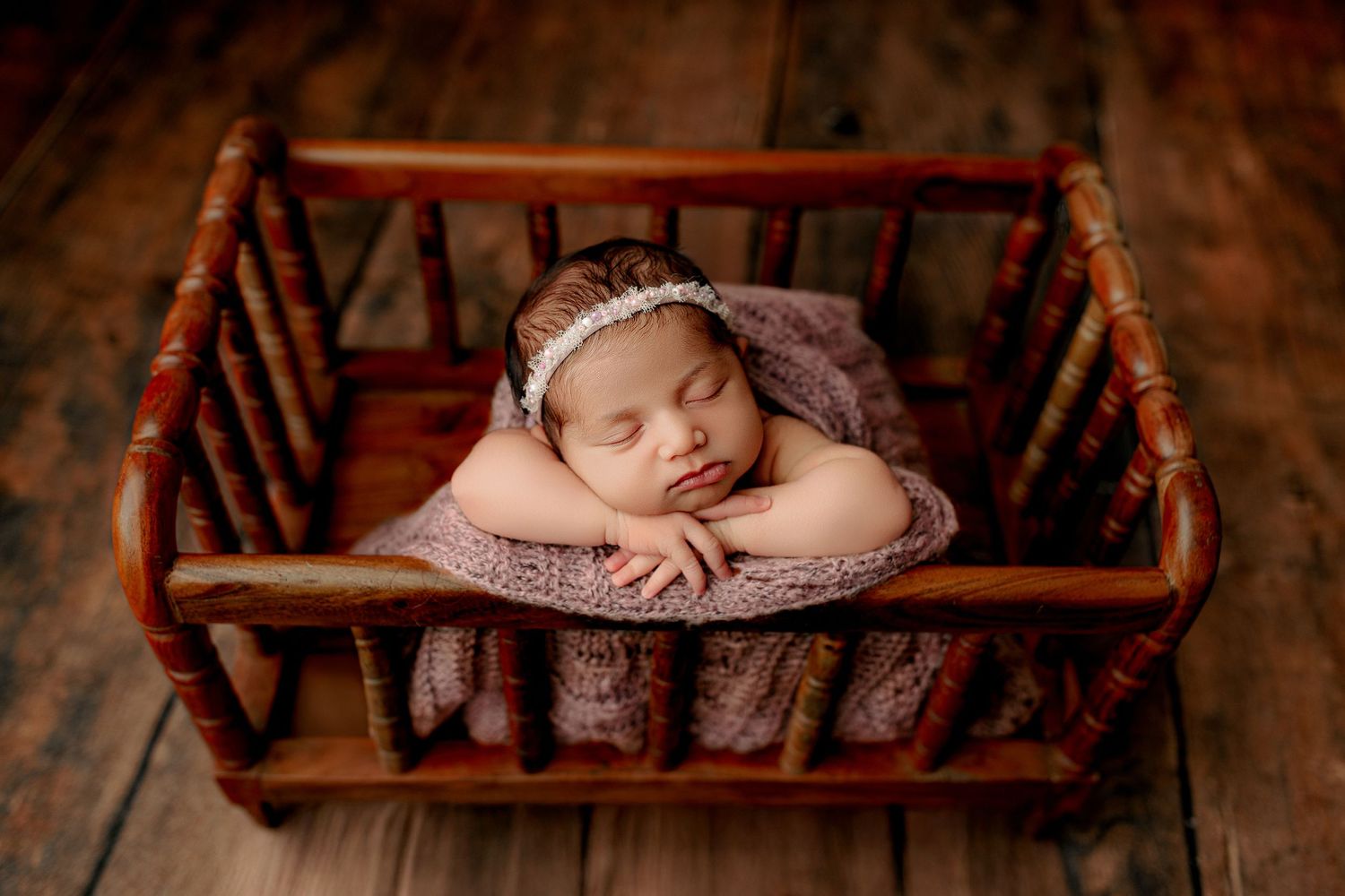 A sleeping baby with a headband rests peacefully on a wooden bed, covered with a purple knitted blanket. The scene is warm and serene
