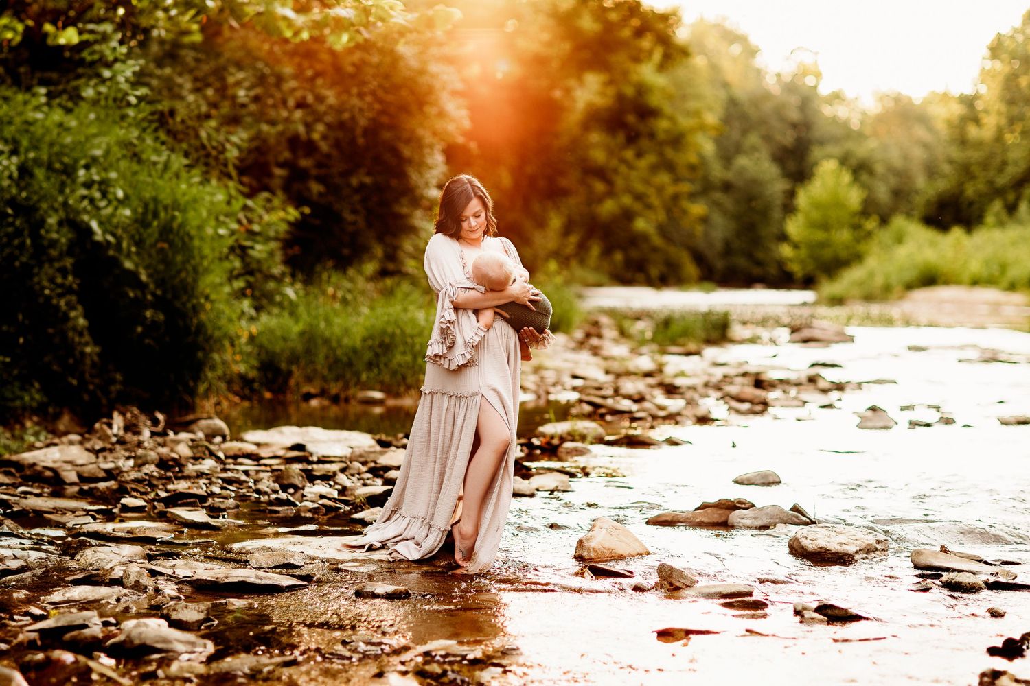 A woman in a flowing dress holds a baby while standing on a rocky riverbank. Sunlight filters through the trees, creating a serene scene