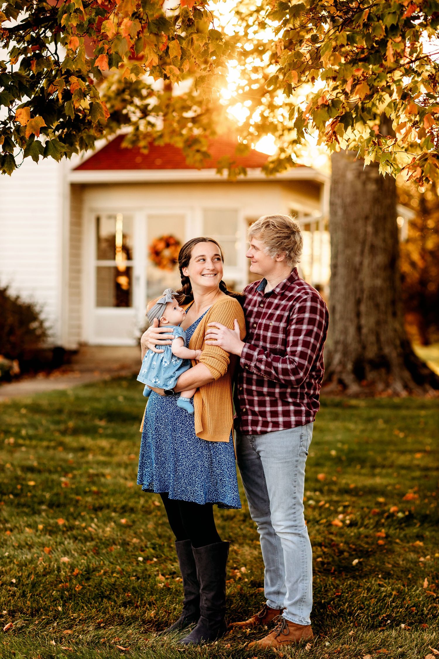A couple stands in a sunlit yard with autumn leaves. The woman holds a baby, and they share a warm smile. A cozy house is in the background.