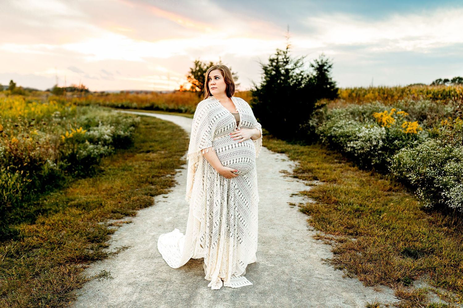 A pregnant woman in a flowing white lace dress stands on a path amid fields at sunset, hand resting on her belly, conveying serenity