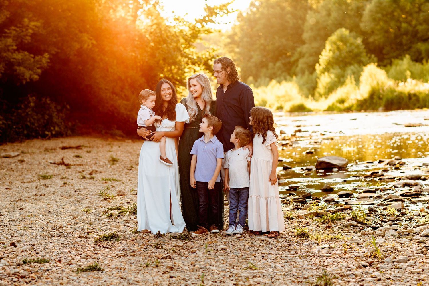 Family portrait by a sunlit stream. Six people, including children, stand on a pebbled shore. Warm, golden sunlight and lush greenery