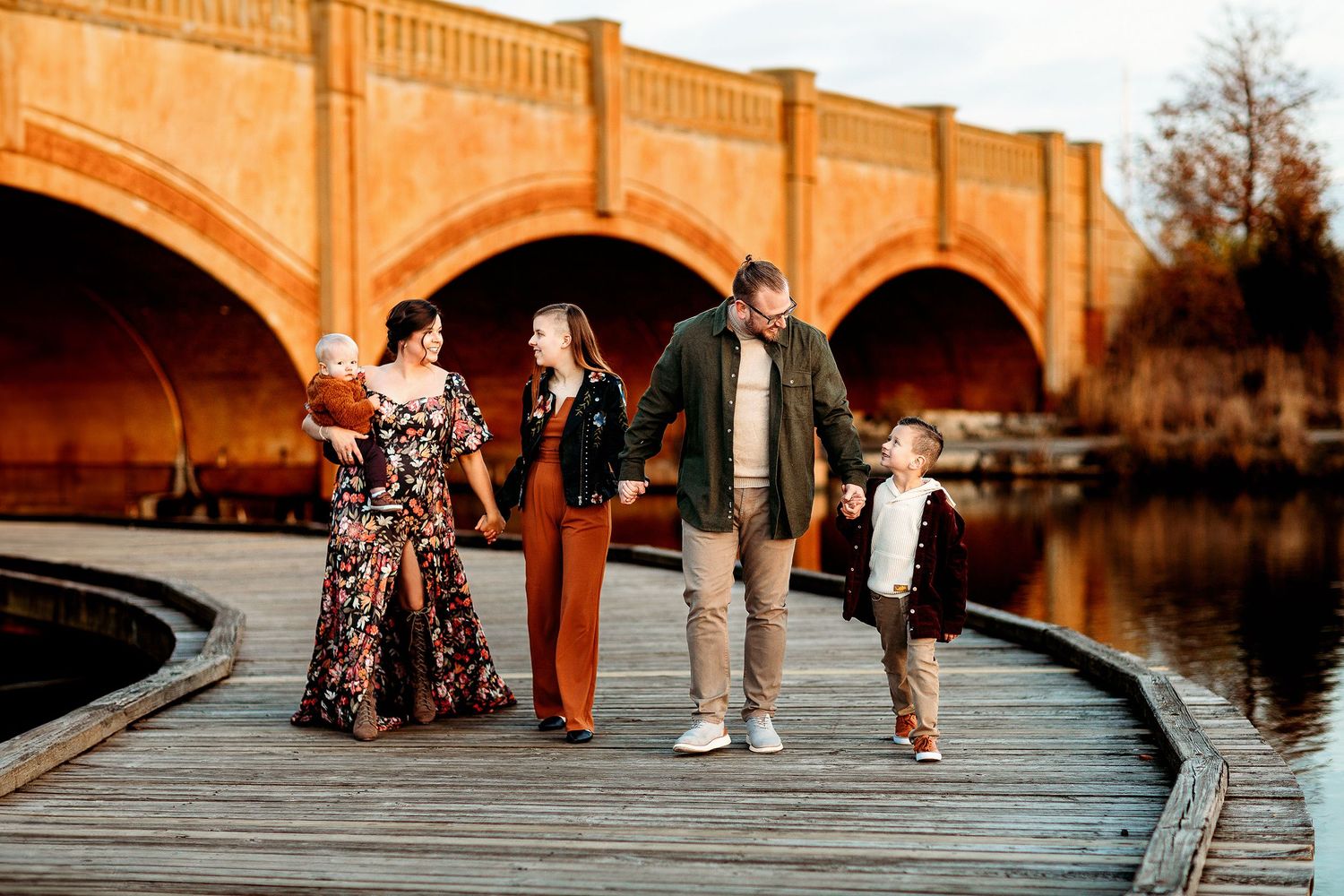 Family of five walks hand in hand on a wooden boardwalk near an arched brick bridge, sharing smiles and joy.