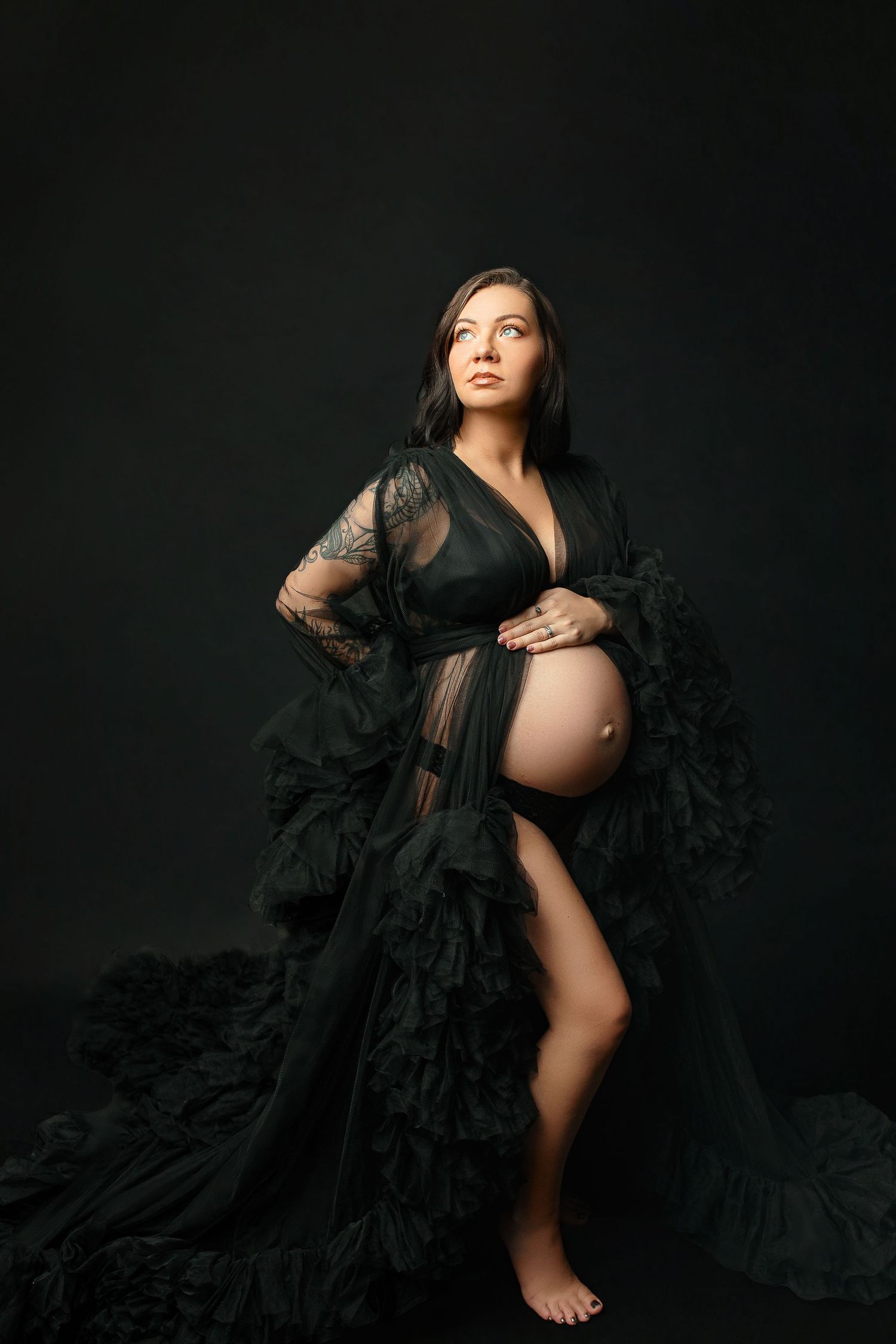 Pregnant woman posing confidently against a dark background, wearing a flowing black sheer gown that reveals her belly.