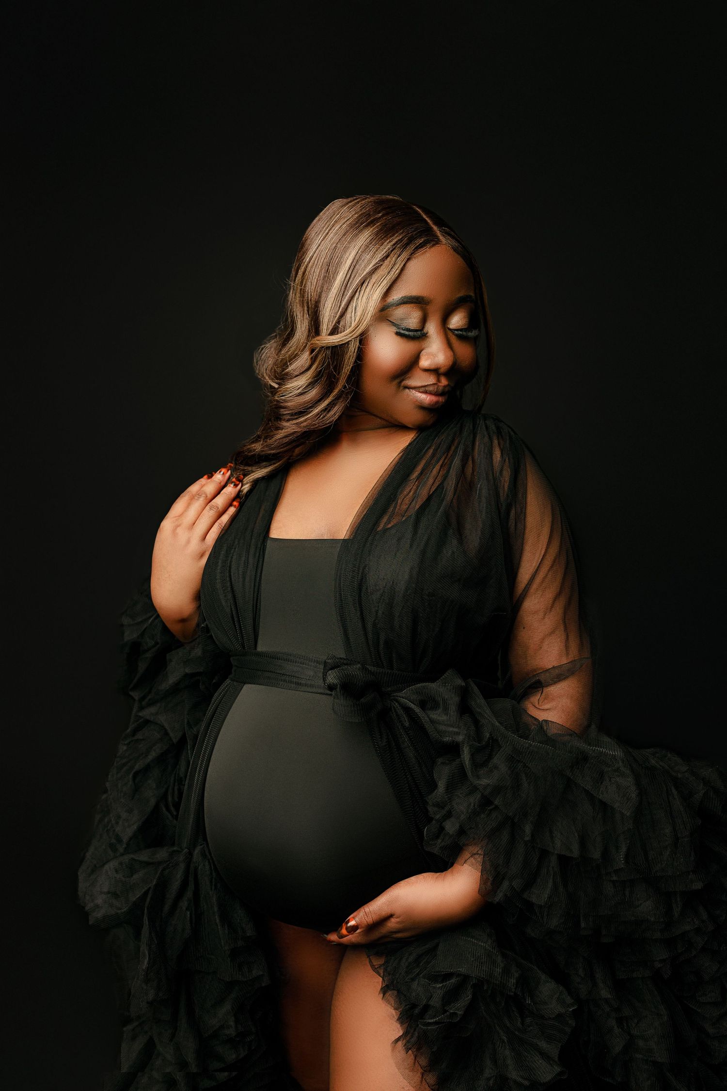 Pregnant woman in a flowing black dress cradles her belly, smiling gently with closed eyes. The dark background accentuates her joy