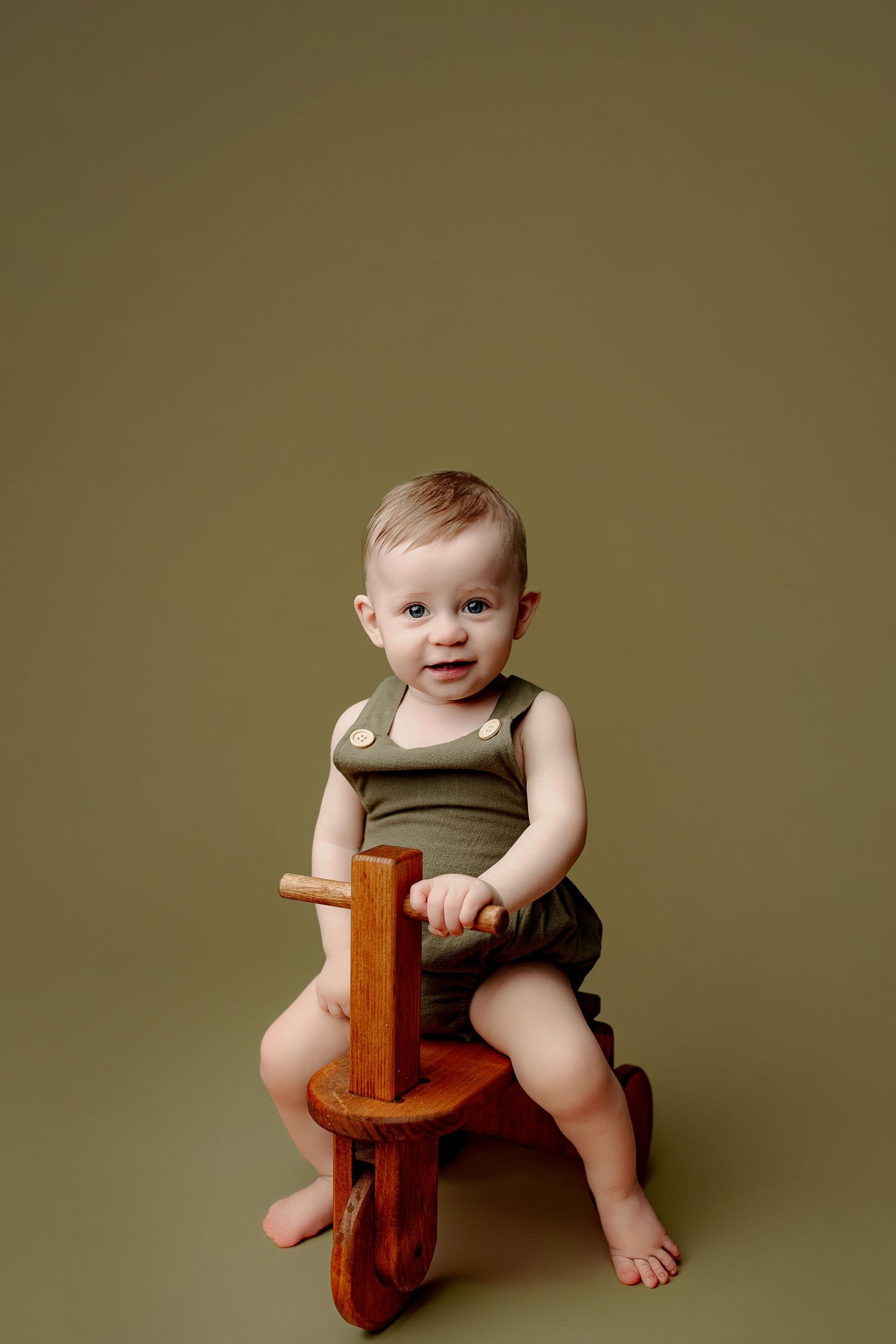 A smiling baby in a green outfit sits on a wooden toy horse against green background for a timeless baby photo Indianapolis Indiana