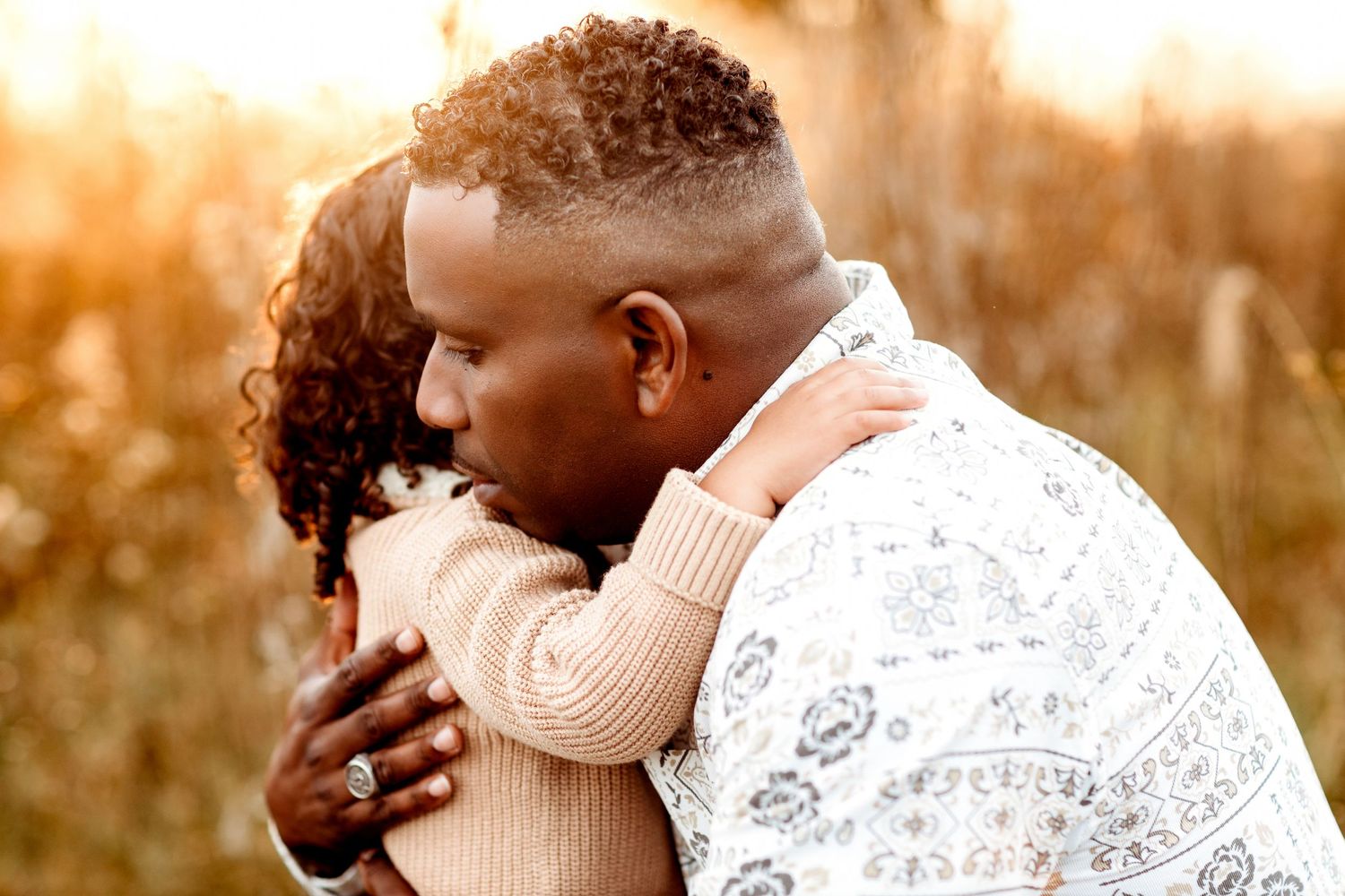 A man embraces a child in a warm hug outdoors, bathed in golden sunlight. The background is a blurred natural setting