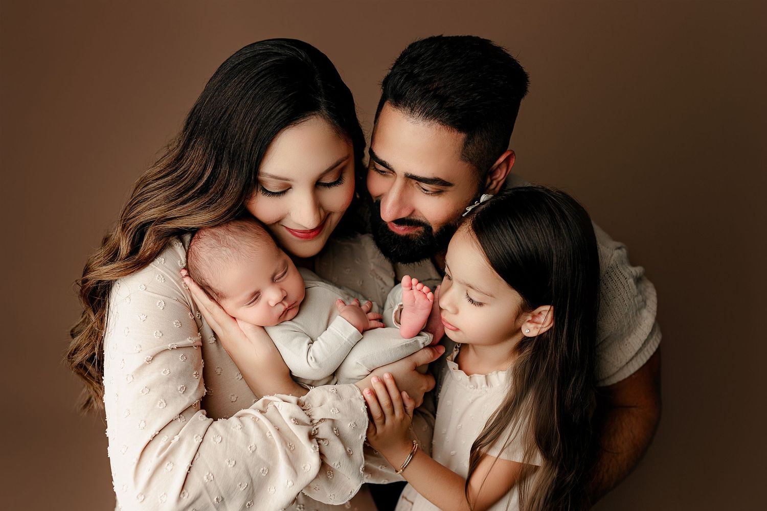 A loving family of four huddles together against a brown background. The mother cradles a newborn, while the father and daughter look lovely