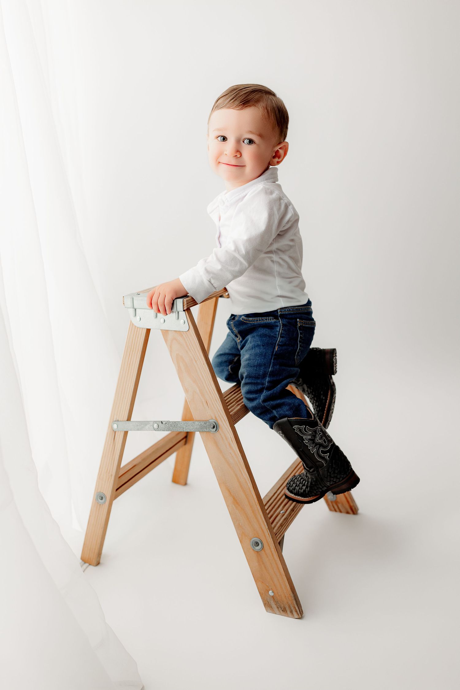 A toddler wearing a white shirt, jeans, and cowboy boots sits playfully on a wooden ladder in timeless Indianapolis baby studio photo