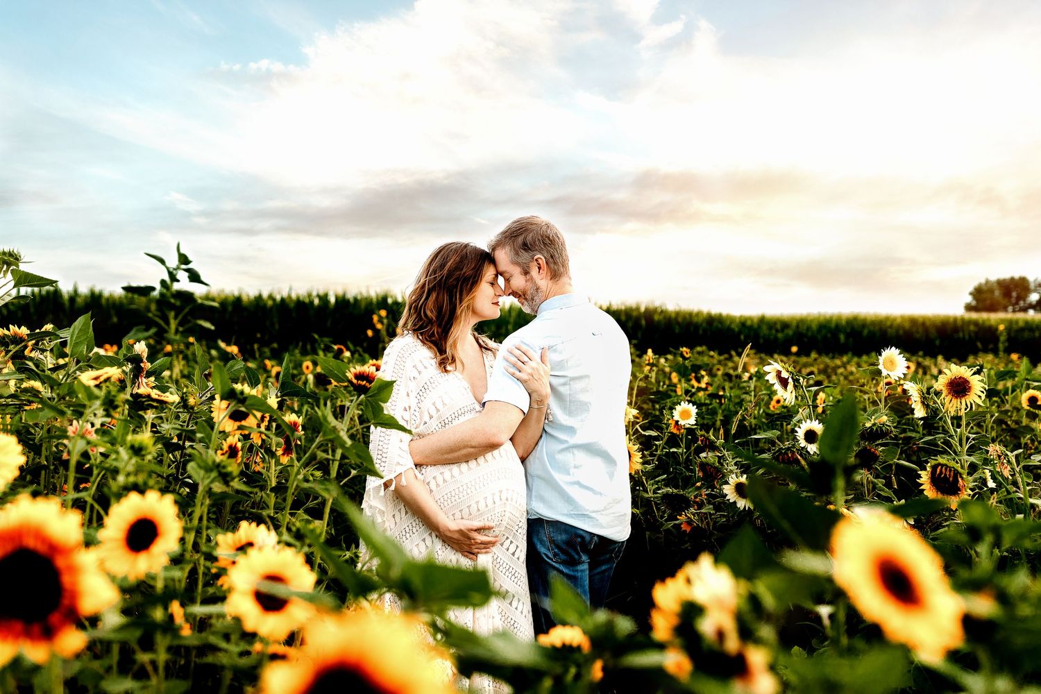 A couple embraces lovingly in a sunflower field under a bright sky. The woman is pregnant, and both are smiling
