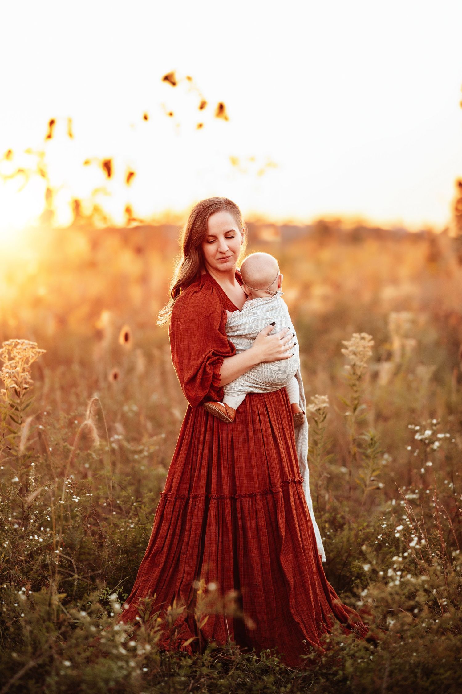 A woman in a flowing red dress holds a baby wrapped in a cloth in a sunlit meadow. Warm tones, wildflowers, and soft light