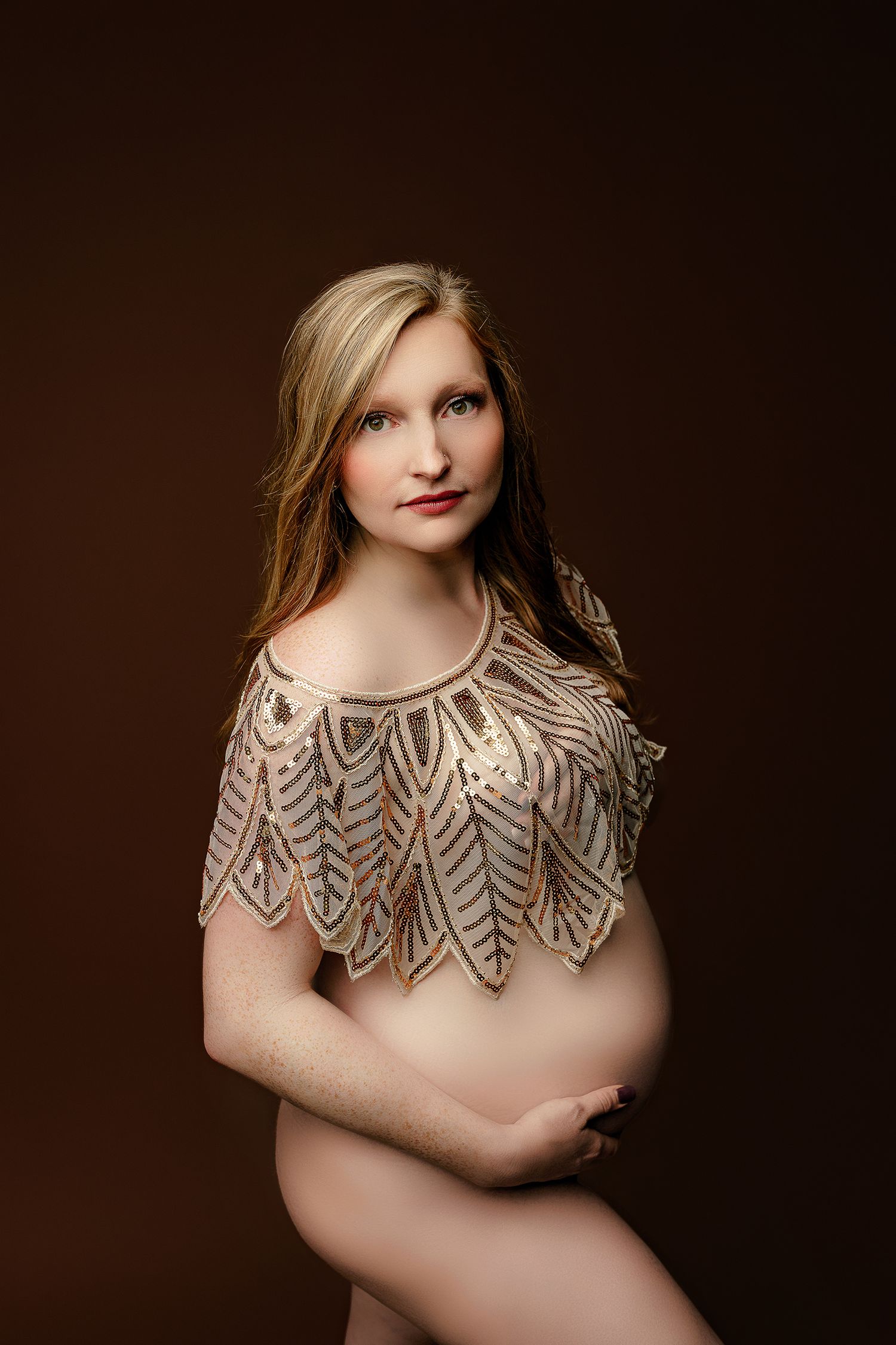 Pregnant woman in a shimmering, decorative top holds her belly against a dark background. Her expression is serene