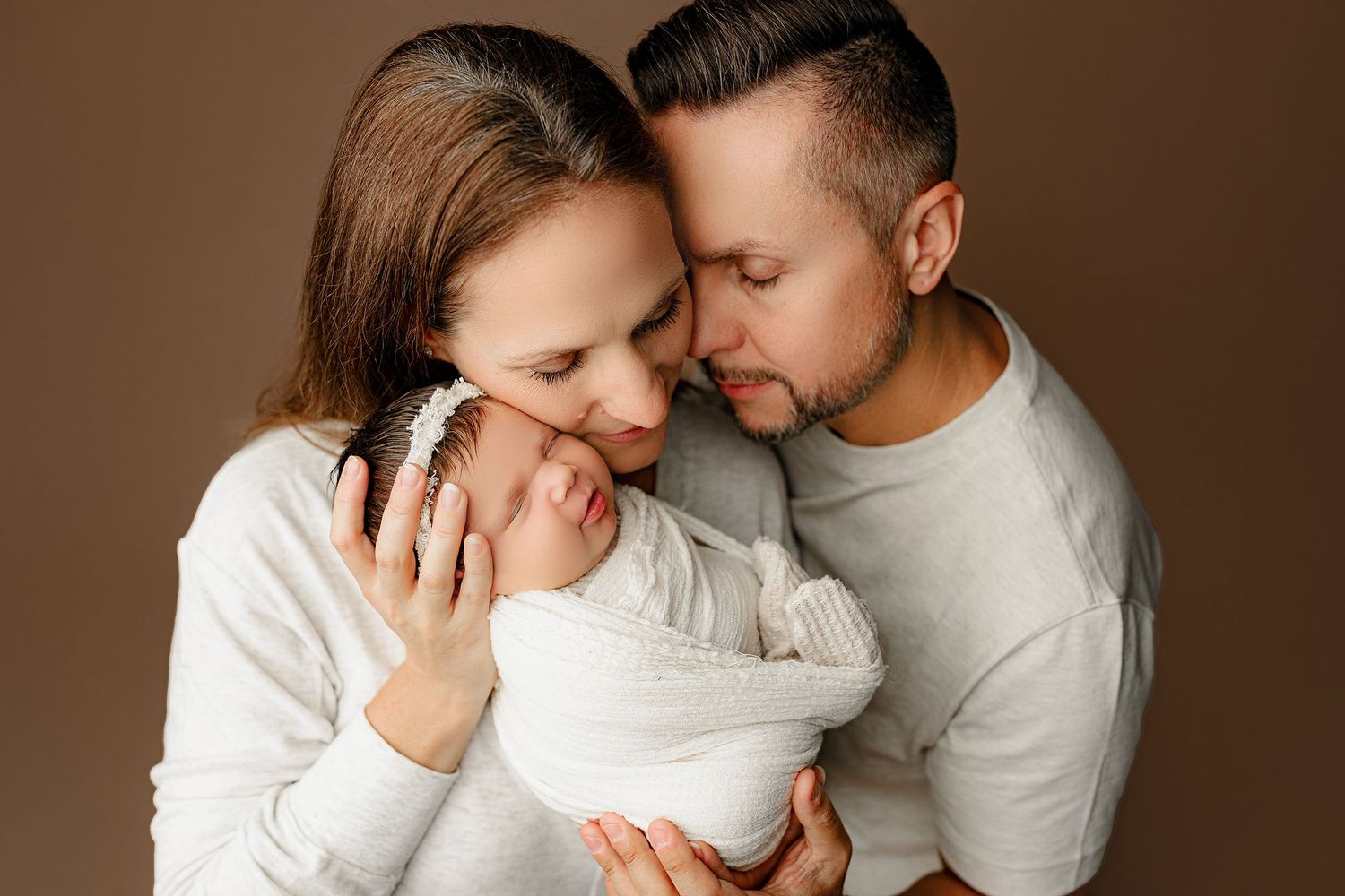 A couple lovingly embraces their newborn, wrapped in a white blanket. The parents gently touch their heads together, conveying love