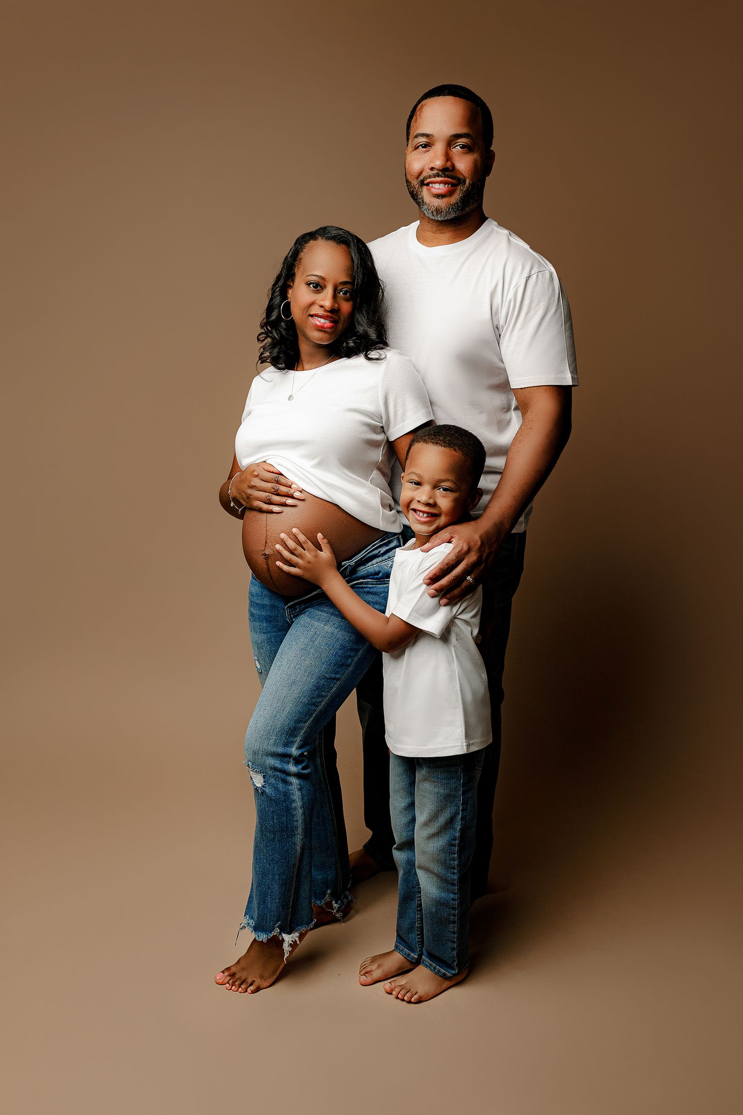 A smiling family poses against a brown backdrop. The pregnant mother and father wear white shirts and jeans, while their son hugs them