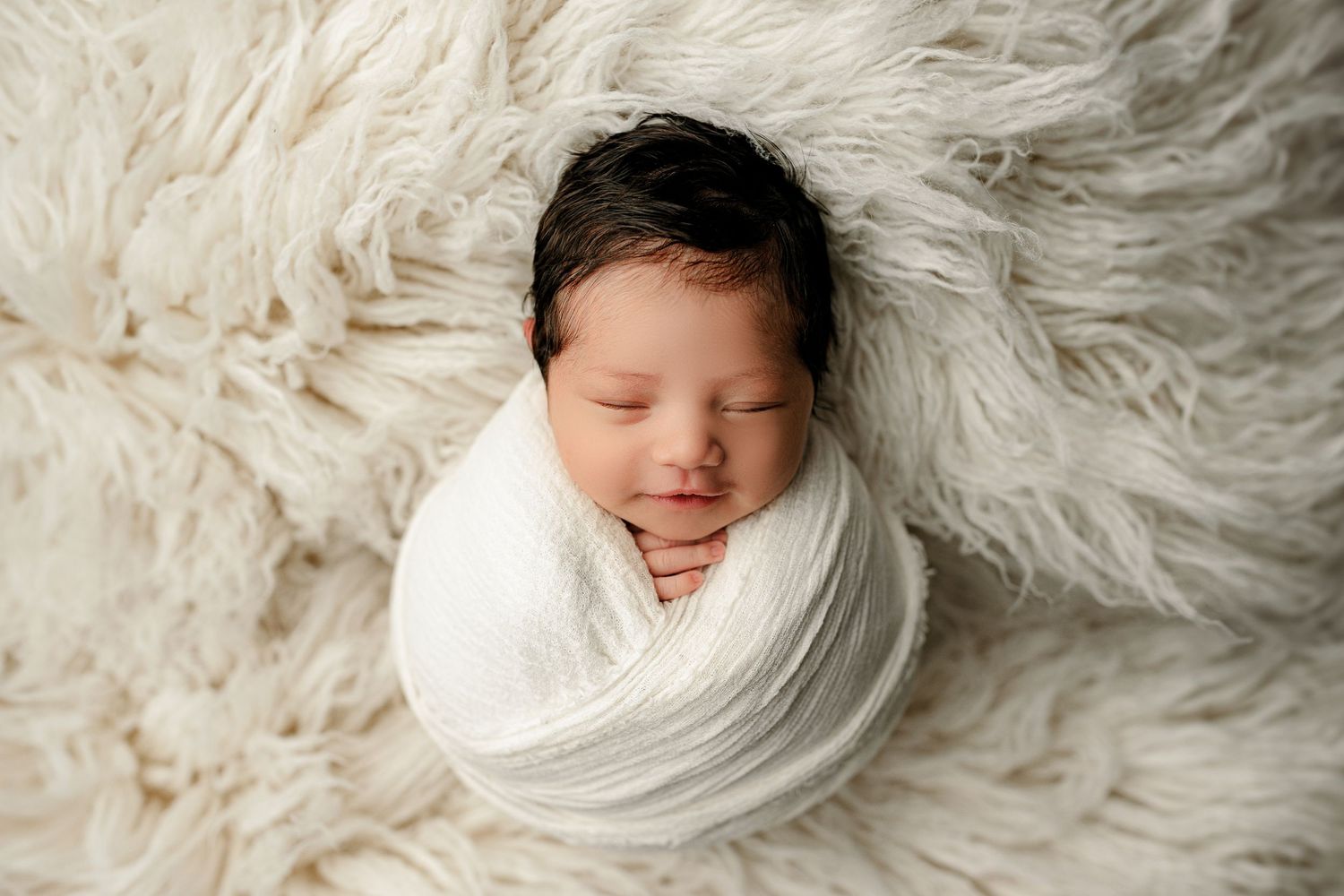 A peacefully sleeping baby with dark hair is snugly wrapped in a white blanket, lying on a fluffy white rug, conveying warmth
