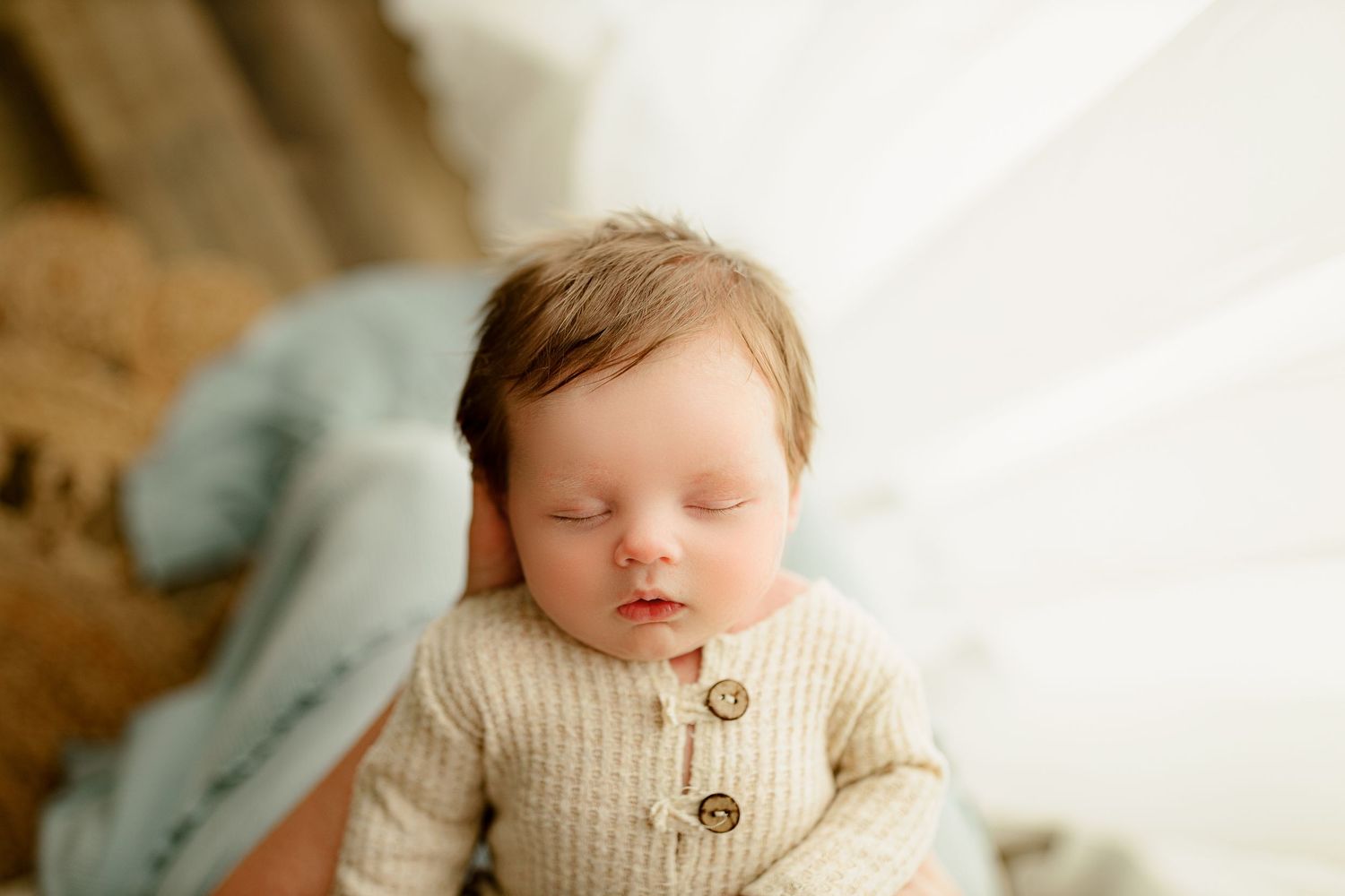 Sleeping baby with brown hair rests peacefully, wearing a cream outfit with buttons. A soft, serene atmosphere is created by gentle lighting