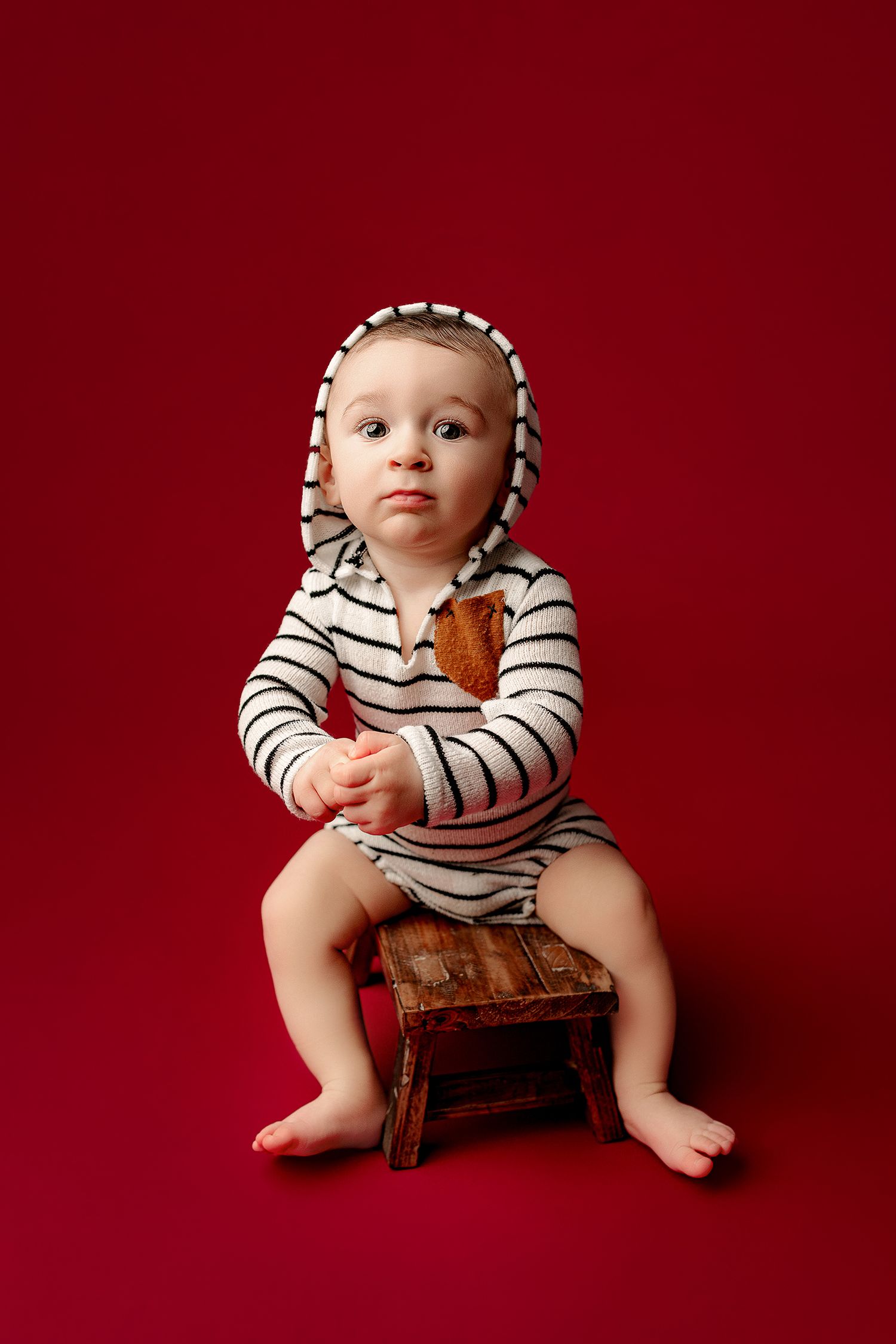 A baby in a striped hoodie sits on a small wooden stool against a deep red background. The atmosphere is playful and inquisitive.