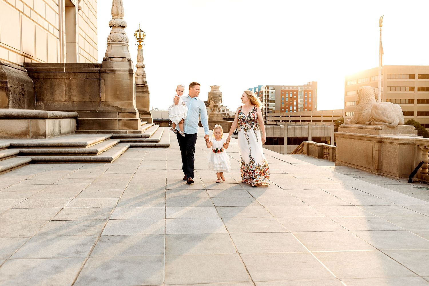 A family of four strolls on a sunlit urban terrace. Parents hold hands with their two children, exuding happiness.