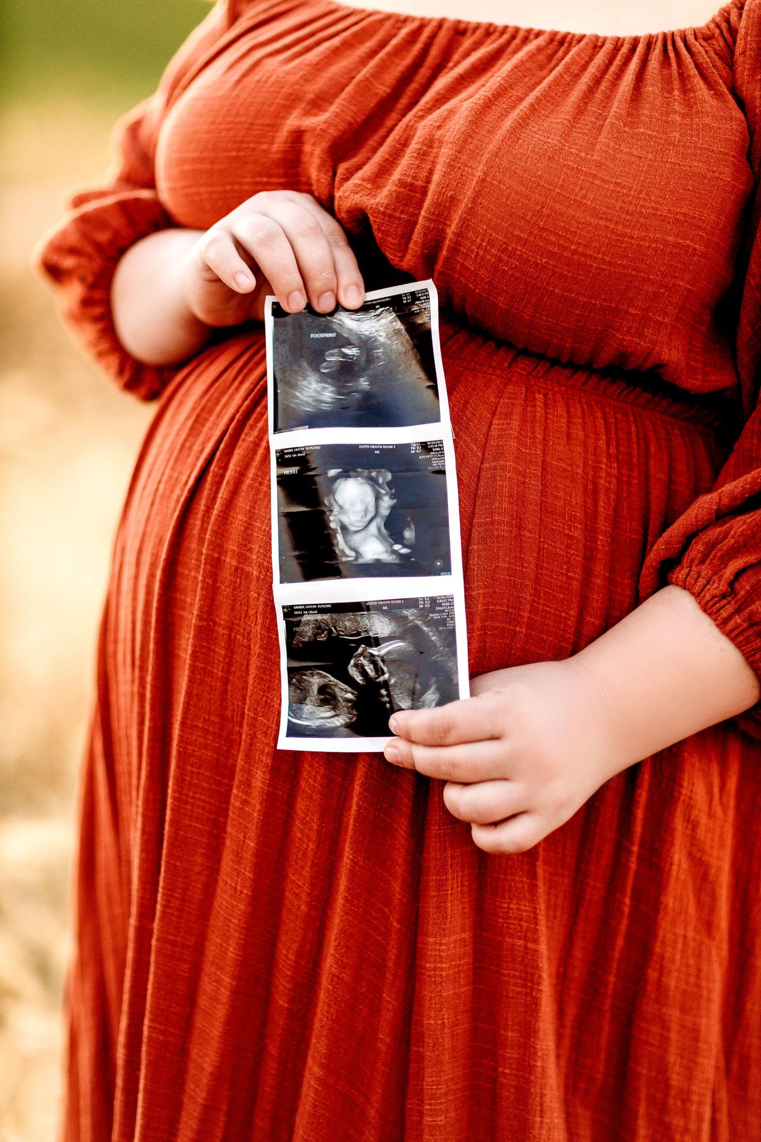 A pregnant woman in an orange dress holds ultrasound images over her belly. The background is softly blurred, creating a warm expectant mood