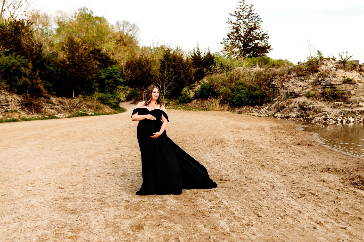 A pregnant woman in a flowing black dress stands on a sandy beach, surrounded by lush greenery, under a bright, cloudy sky