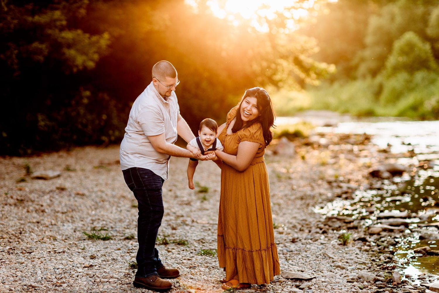 A couple joyfully holds their baby between them on a sunlit, pebbled path near a lush forest. The golden light creates a happy atmosphere