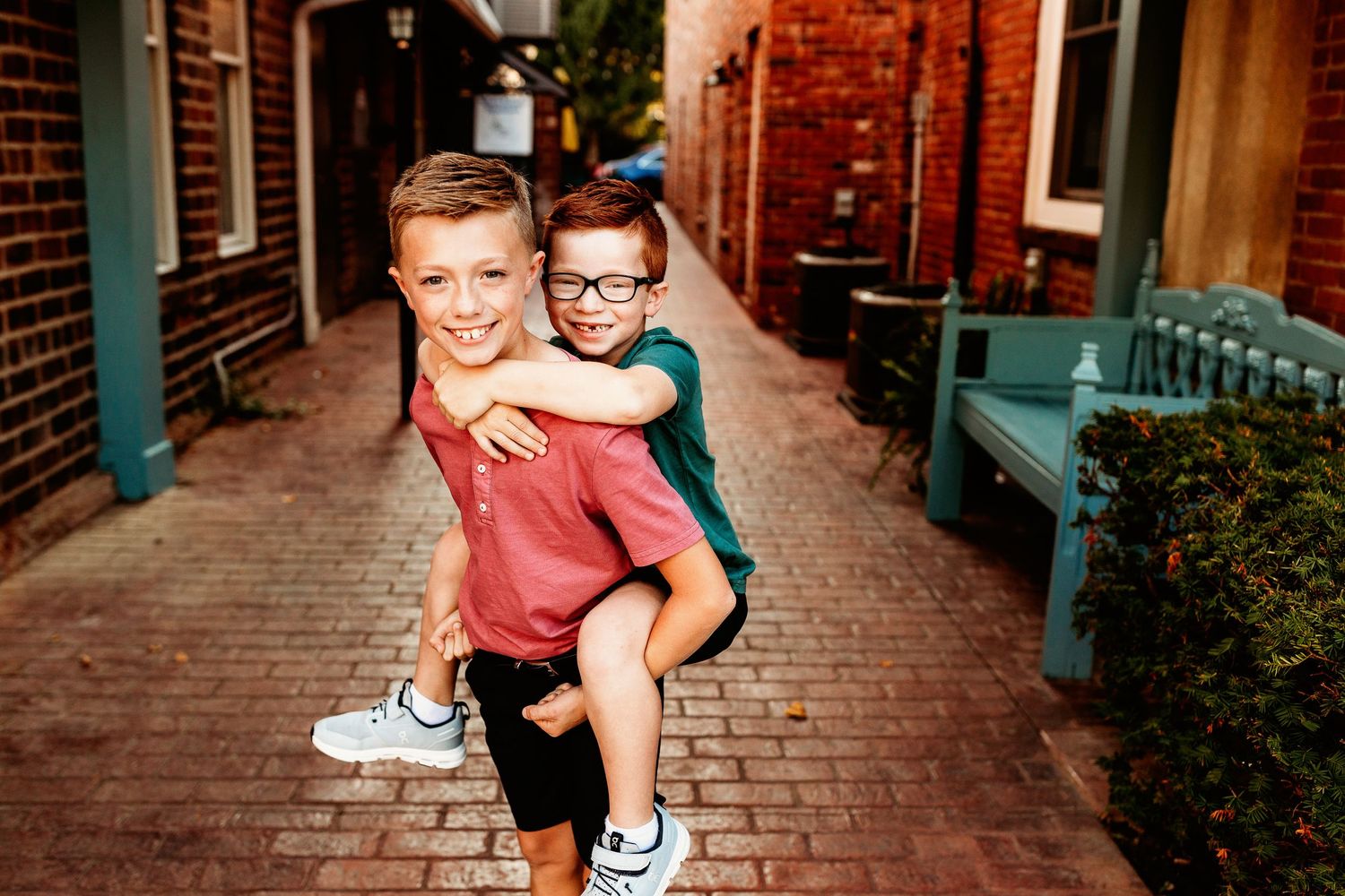 Two smiling boys in casual clothes enjoy a piggyback ride in a narrow, brick-lined alleyway in Zionsville Indiana