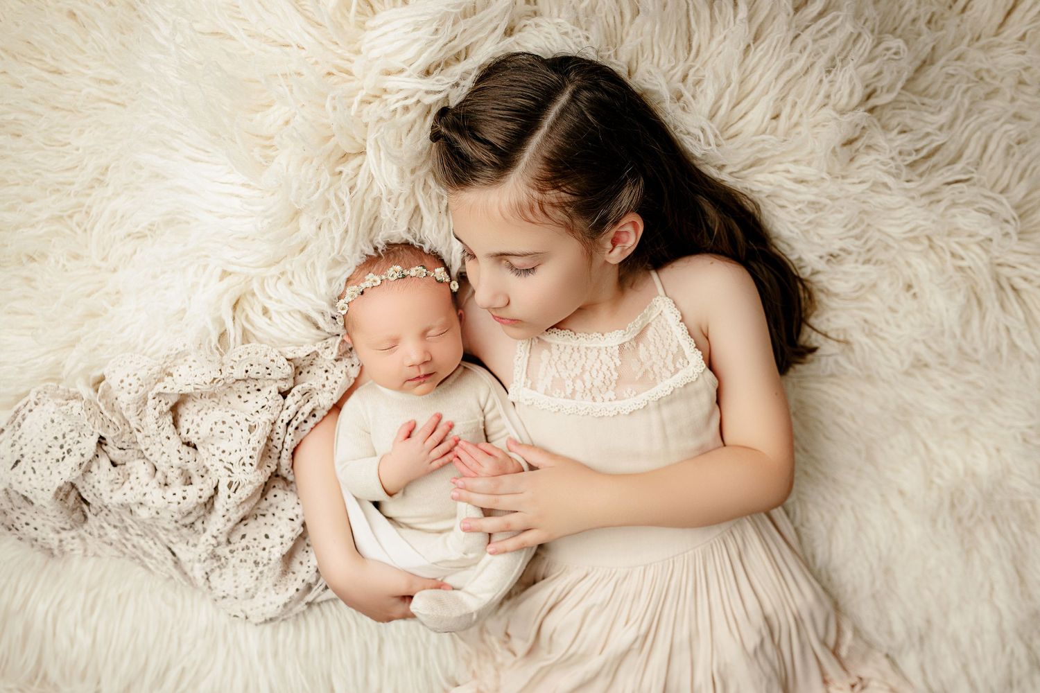 A young girl in a cream dress gently cradles a sleeping newborn in white, both lying on a fluffy blanket. The scene conveys warmth