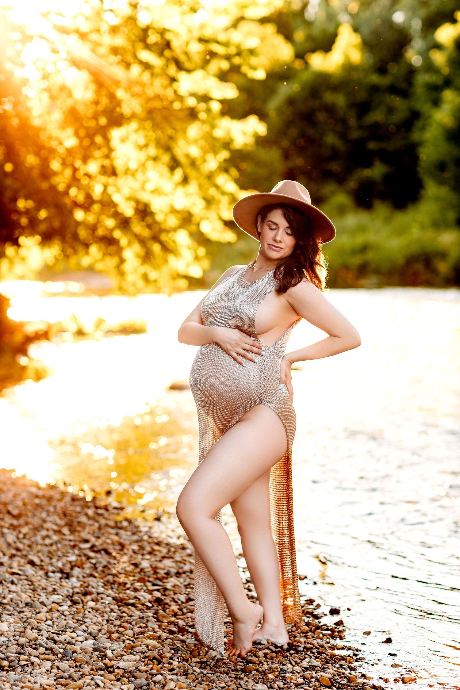 Pregnant woman in a sheer dress and hat stands by a sunlit creek, touching her belly